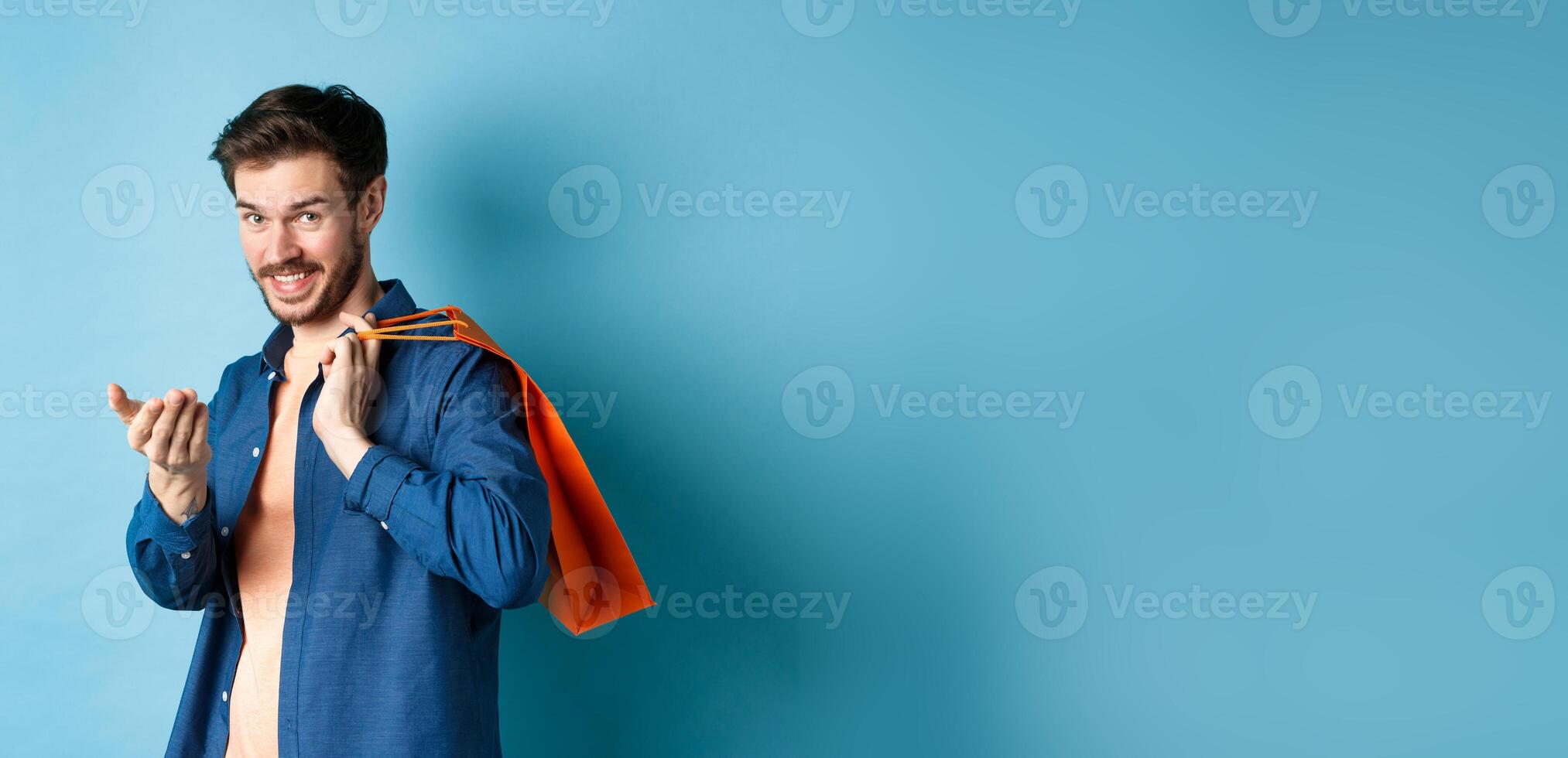 Modern guy holding shopping bag behind shoulder and beckon you, telling to come closer or inviting to store, standing on blue background photo