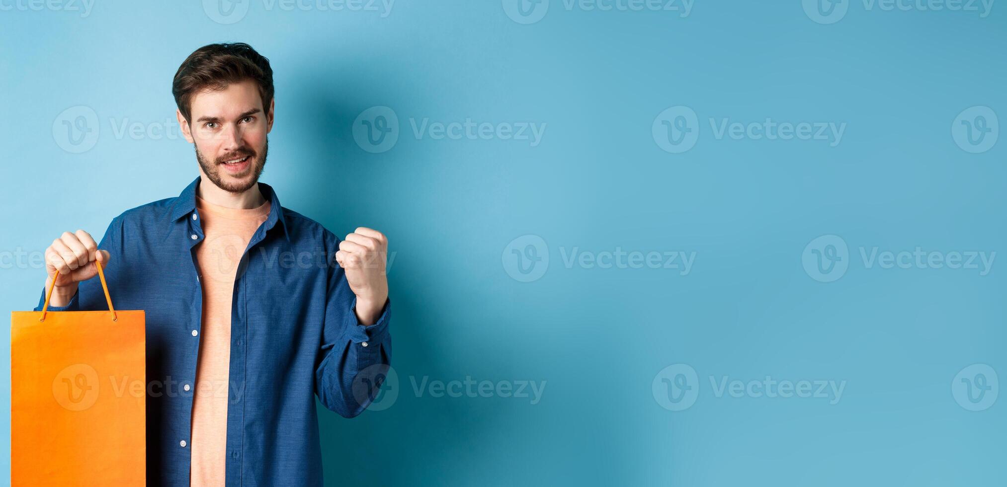 Handsome smiling man saying yes, triumphing and showing shopping bag, celebrate good promo offer, standing on blue background photo