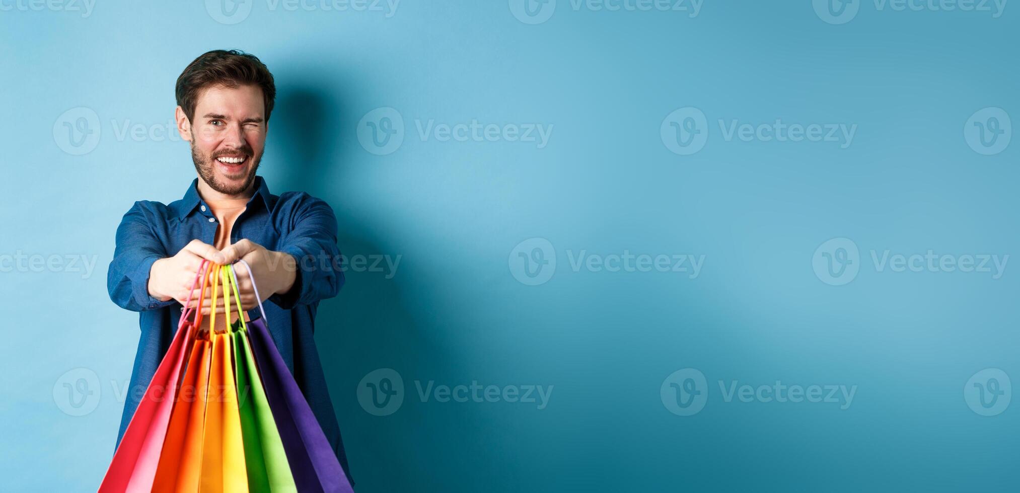 Cheerful handsome man winking and smiling, extending hands with colorful shopping bags, standing on blue background photo