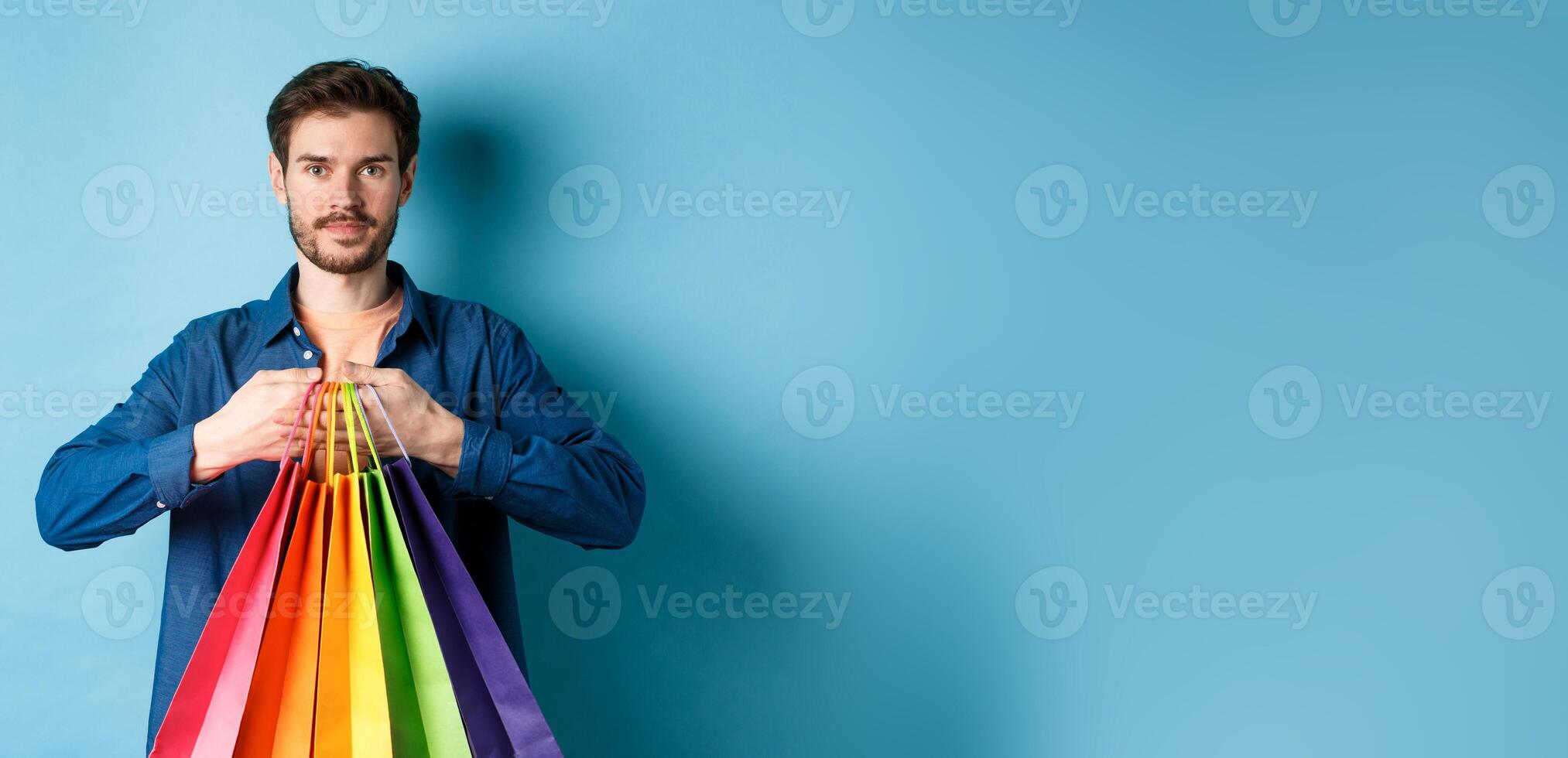 Modern guy with beard, holding packages from shop and smiling, standing on blue background photo