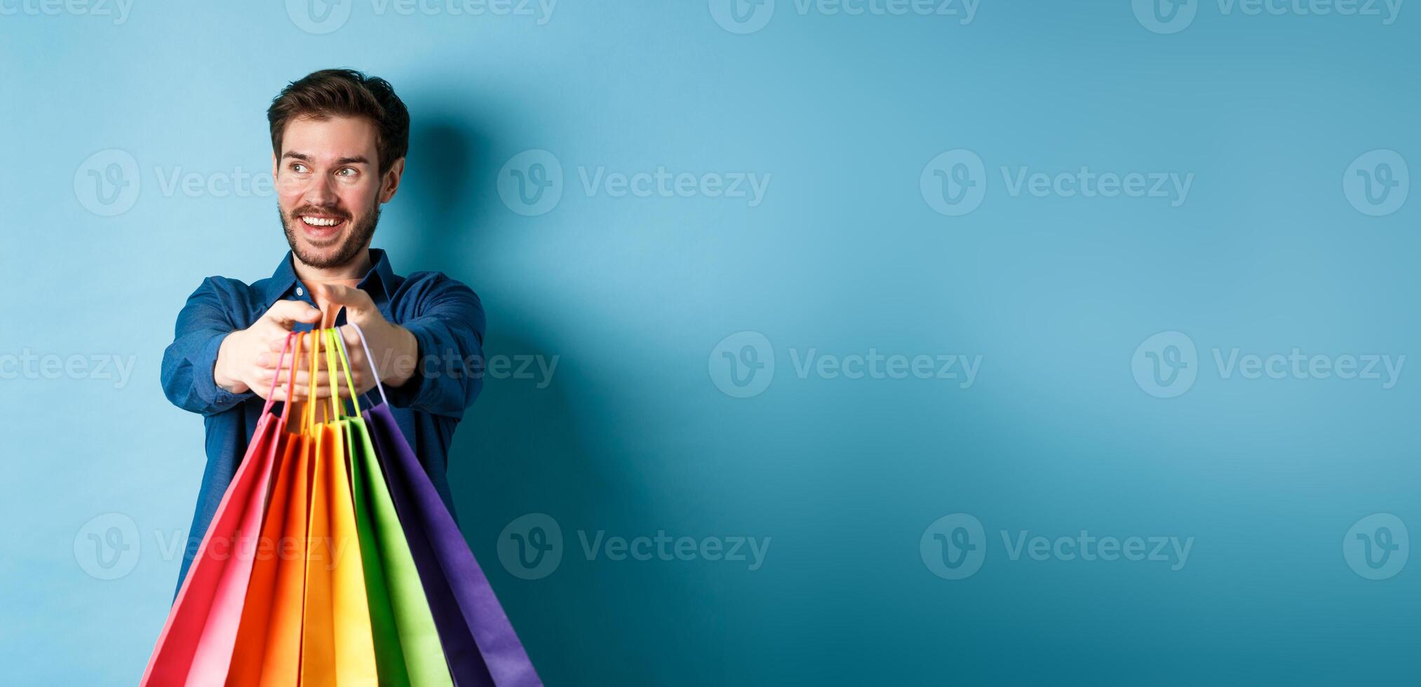 Handsome happy guy smiling, stretch out hands with shopping bags and looking aside at empty space, standing on blue background photo