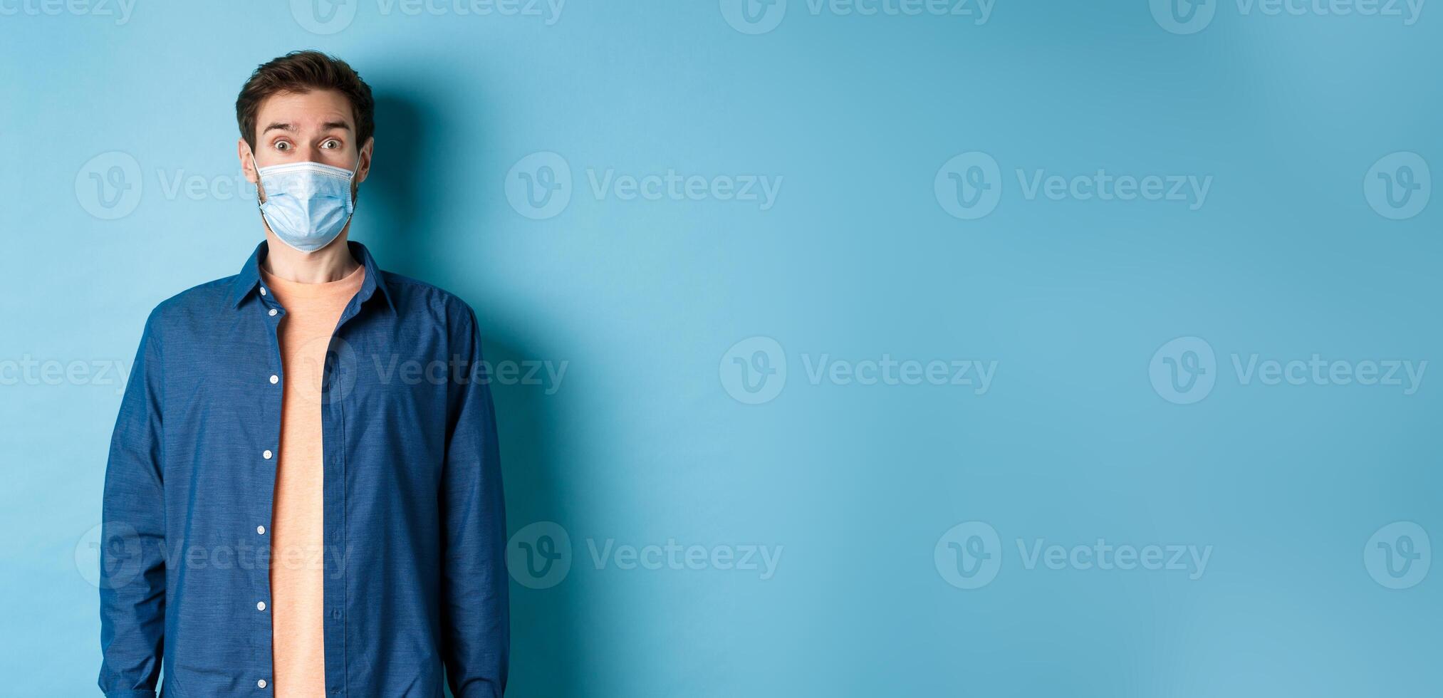 Covid-19 and healthcare concept. Image of handsome modern guy in face mask raising eyebrows in surprise, looking at something in awe, blue background photo