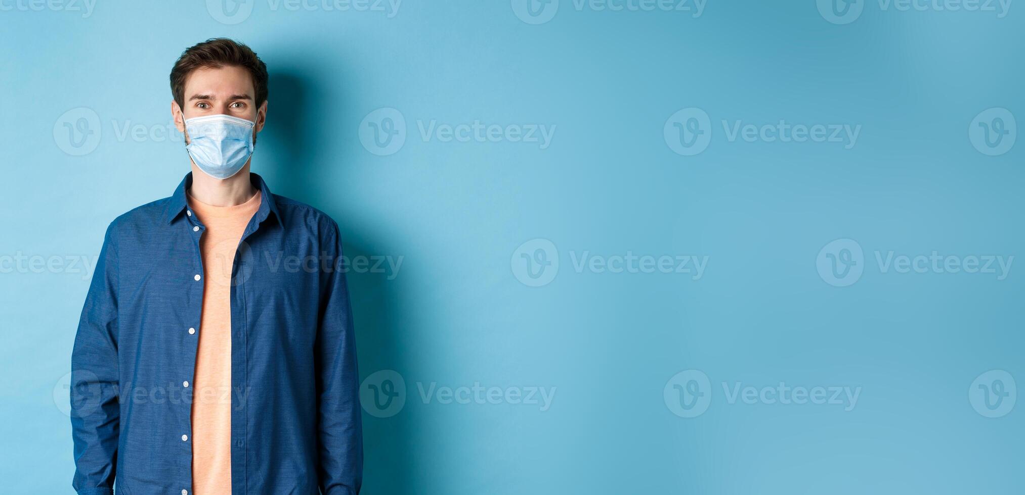Covid-19 and healthcare concept. Smiling young man in medical mask looking healthy and happy, standing on blue background photo