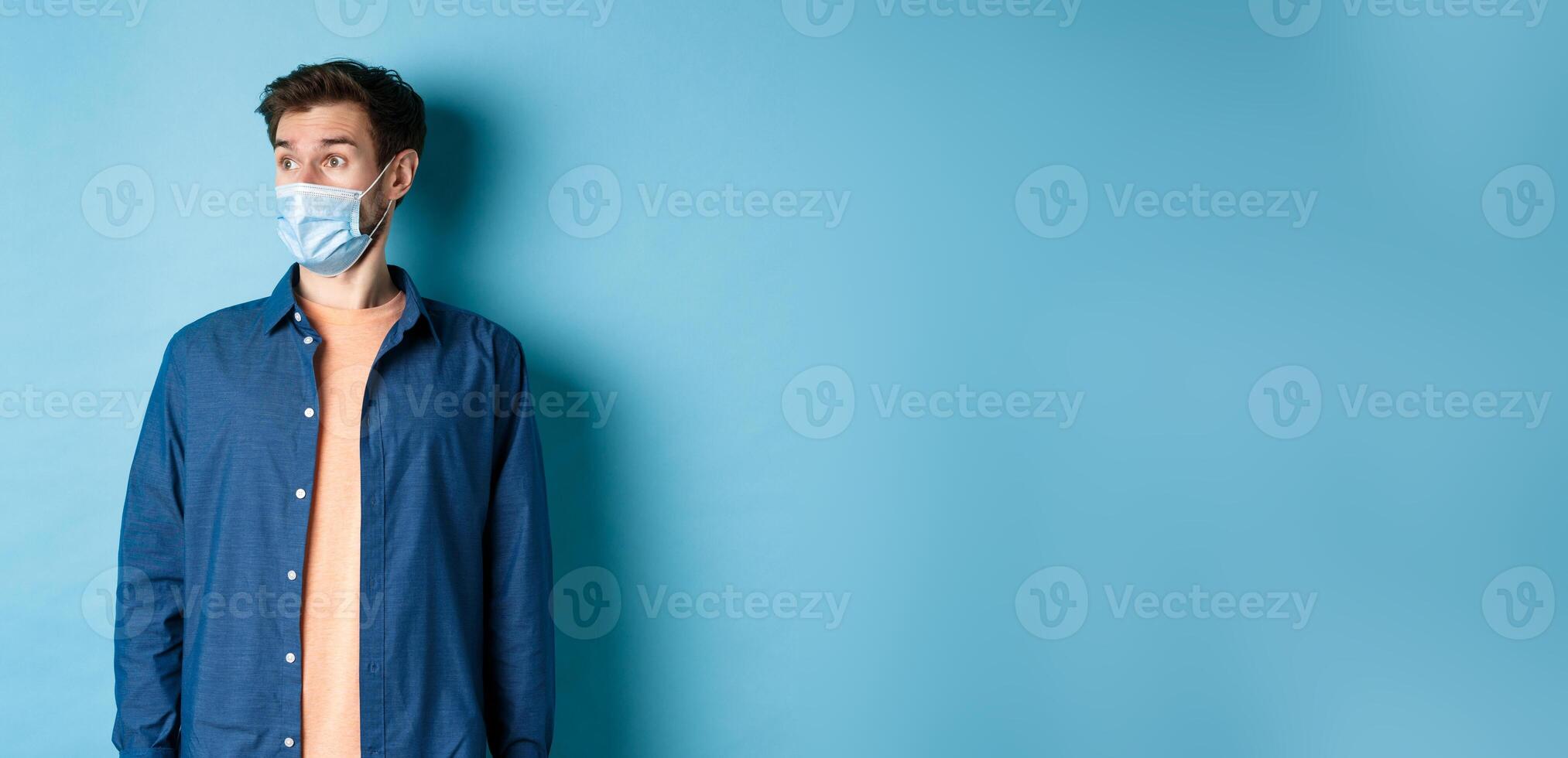 Covid-19 and healthcare concept. Surprised caucasian guy in face mask looking aside at empty space for banner, standing on blue background photo