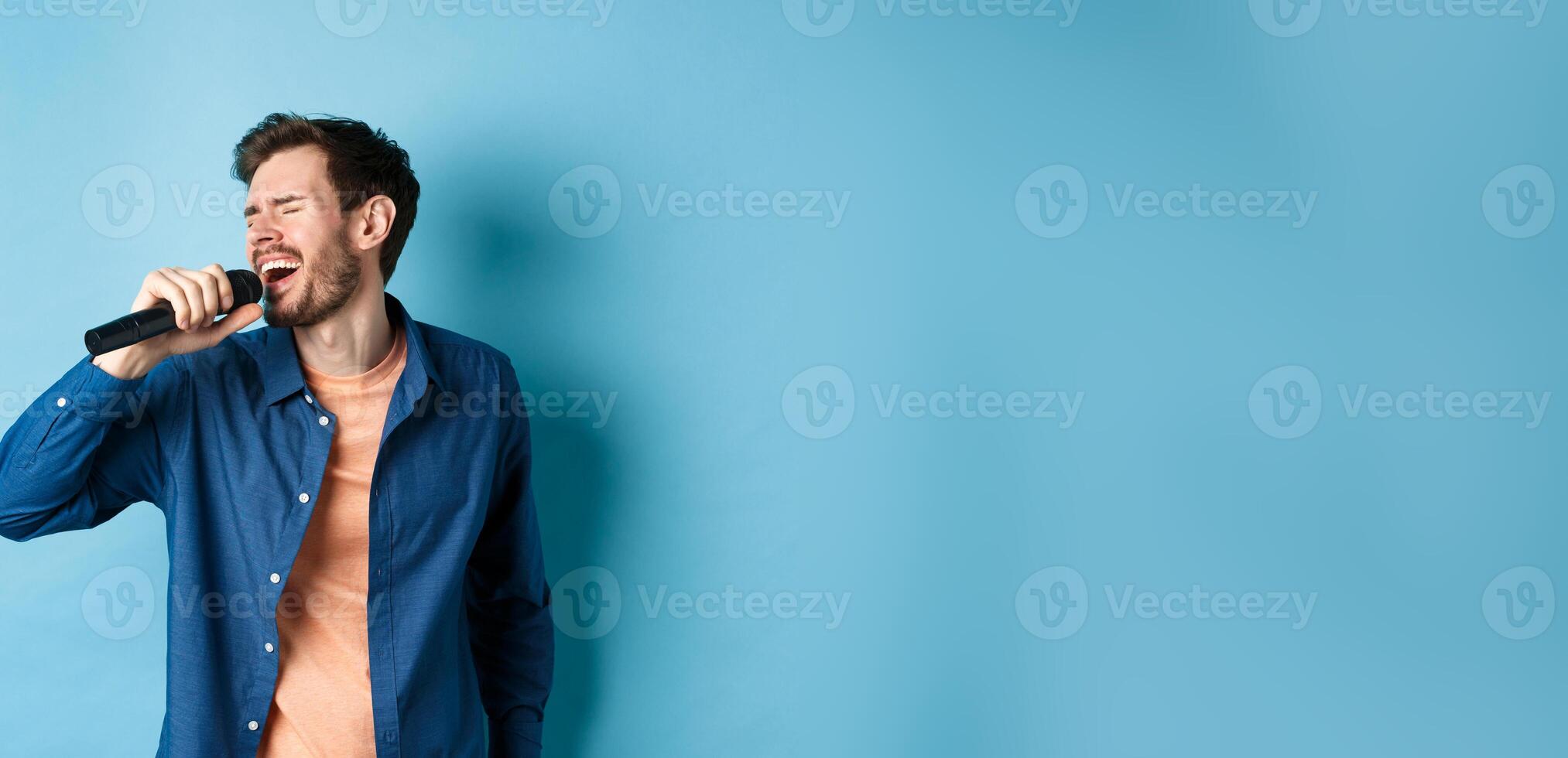 Carefree guy singing karaoke with microphone, perform a song, standing on blue background photo