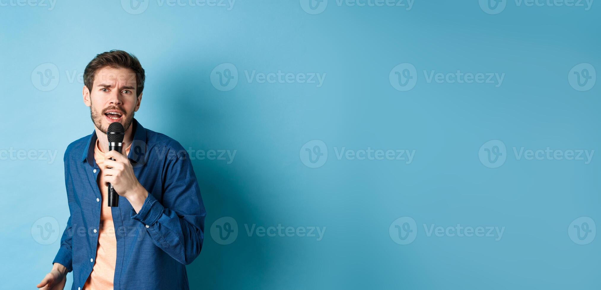 Passionate singer looking at camera, singing in microphone, playing karaoke on blue background photo