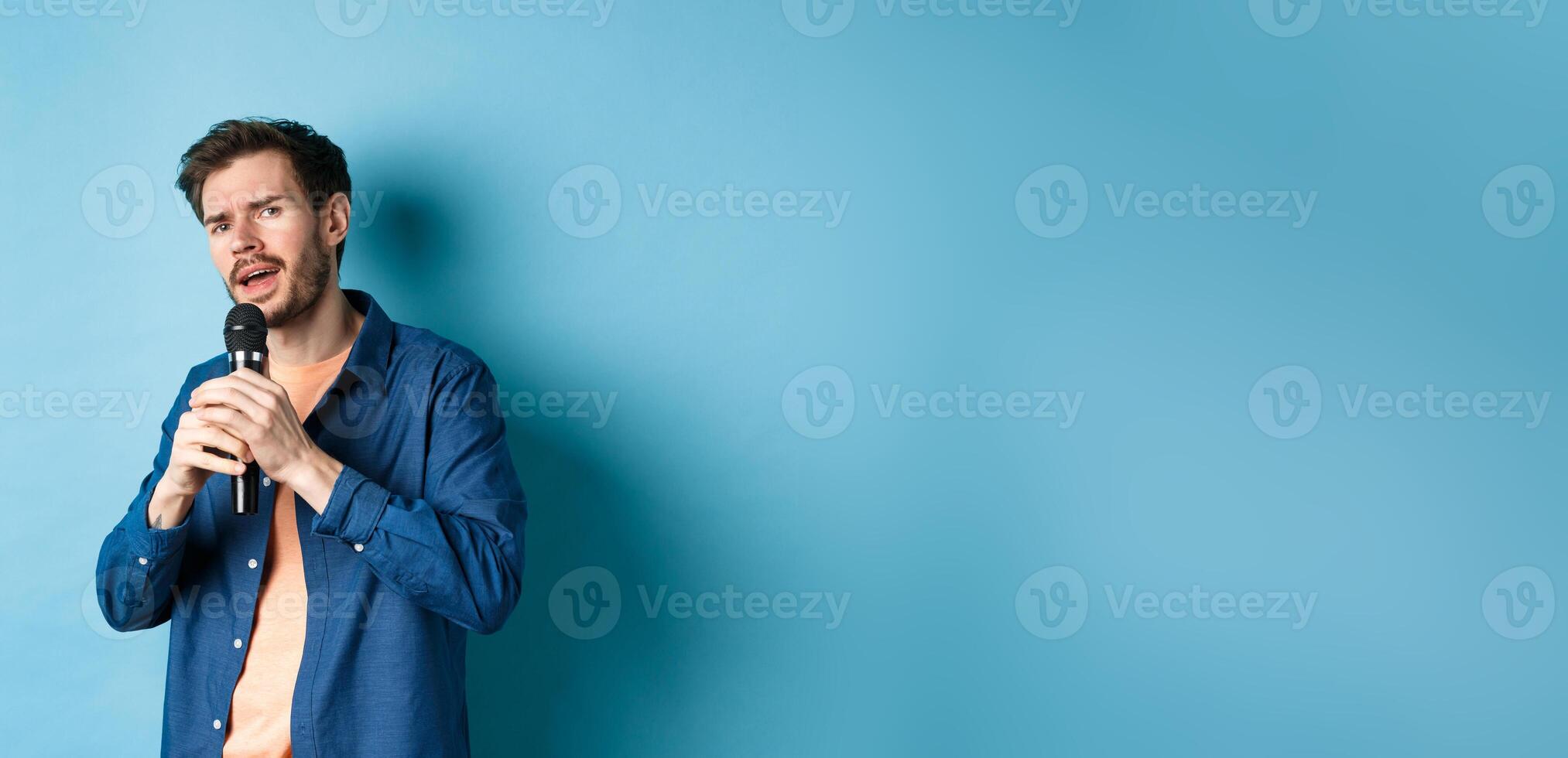 Young man singing song in microphone, playing karaoke, standing on blue background photo