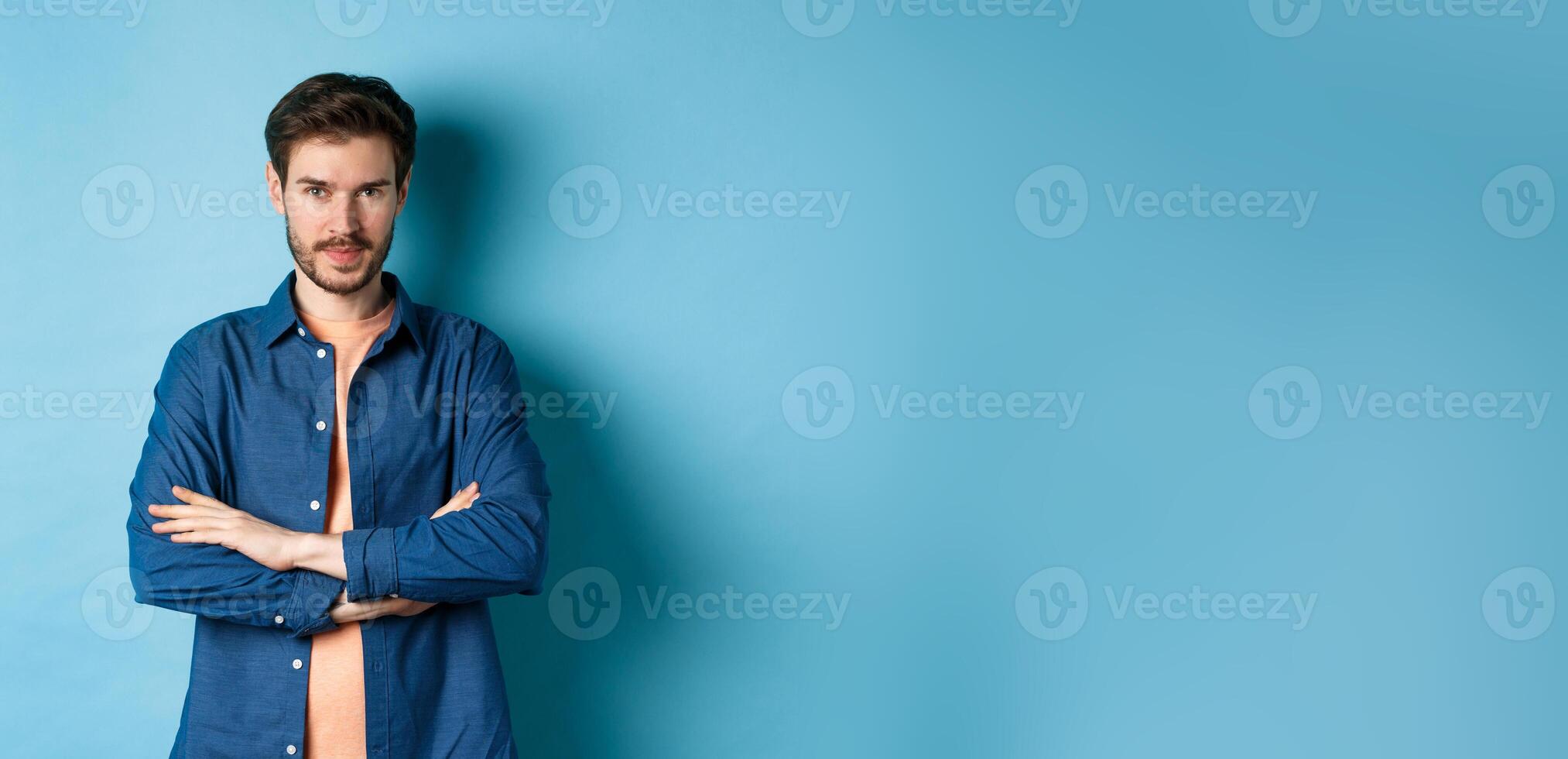 Confident young man feeling like professional, cross arms on chest and smiling sassy, standing on blue background photo