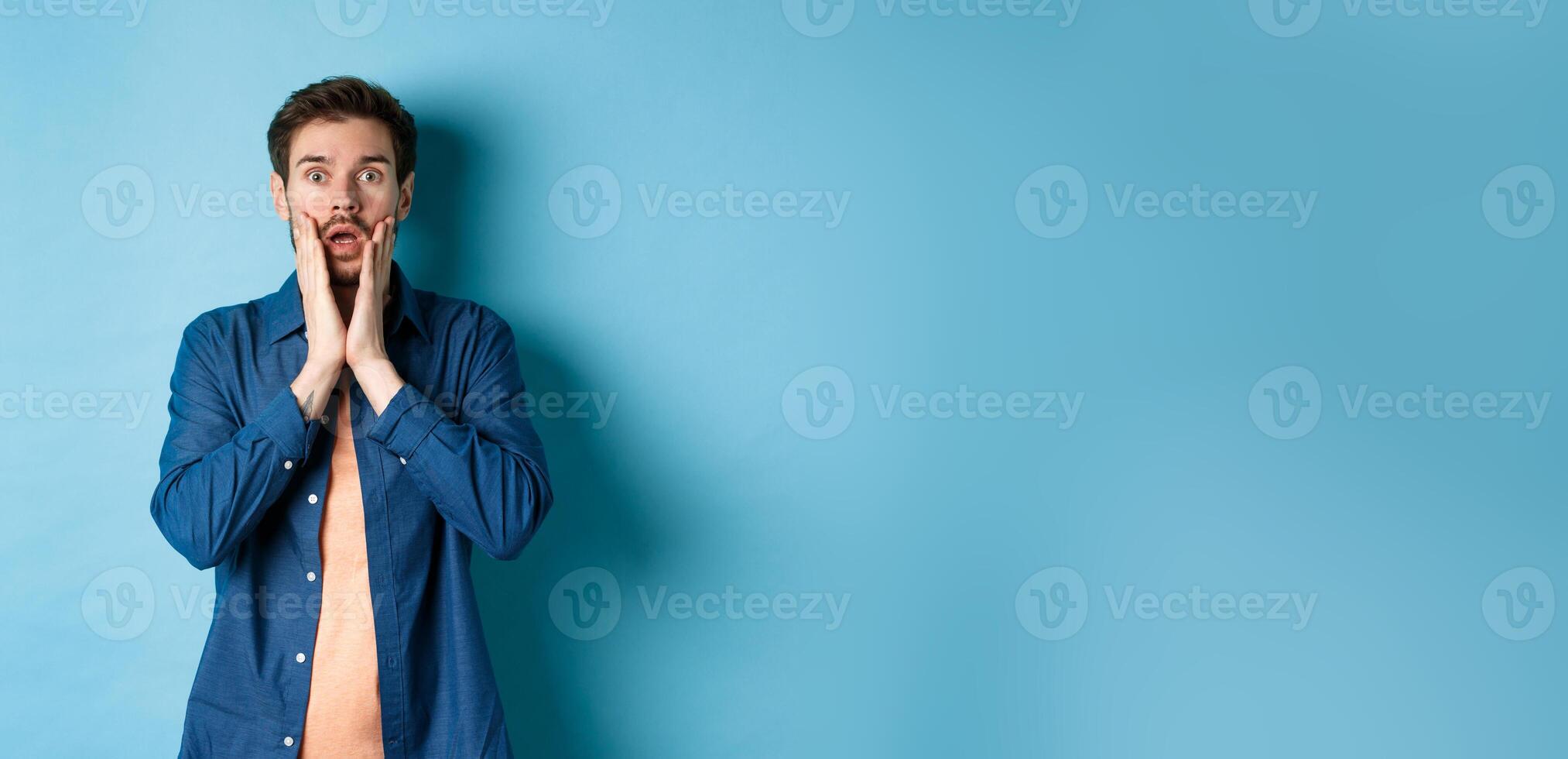 Portrait of guy gasping and dropping jaw with shocked face, staring at camera startled, standing on blue background photo