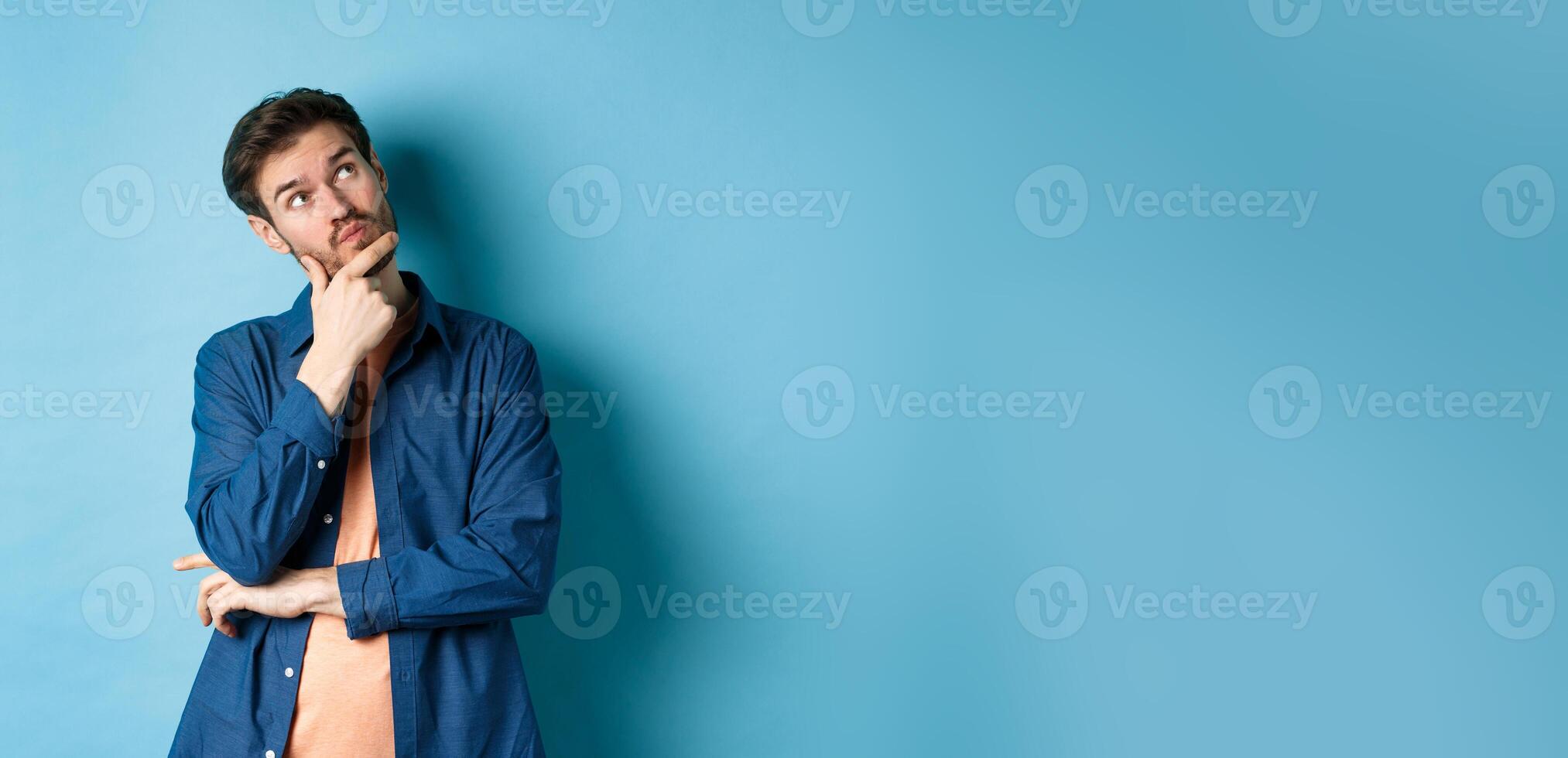 Pensive young man touching face and looking aside at empty space, imaging something, thinking off plan, standing on blue background photo