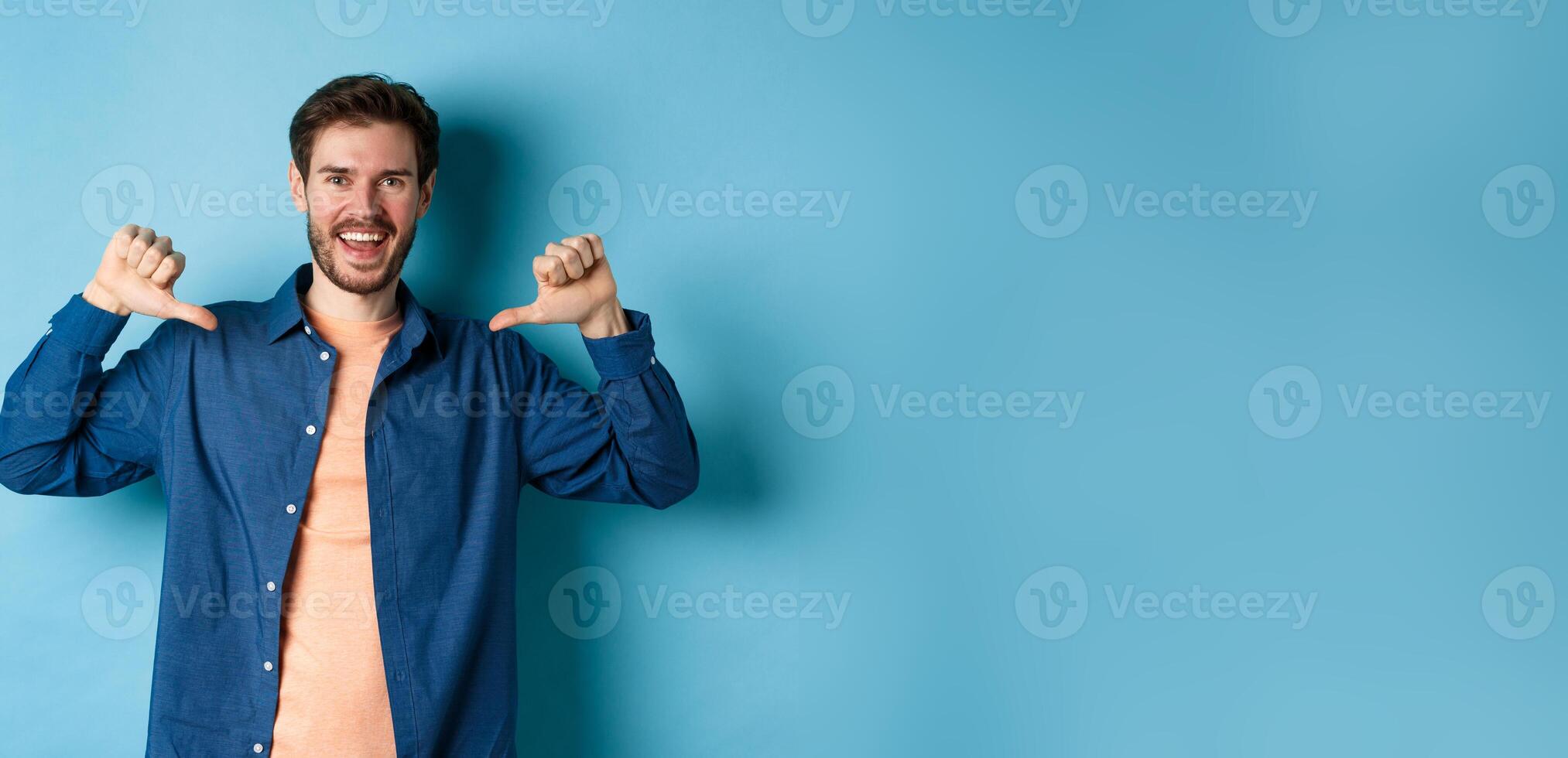 Happy young man self-promoting, pointing at himself and smiling, standing like professional on blue background photo