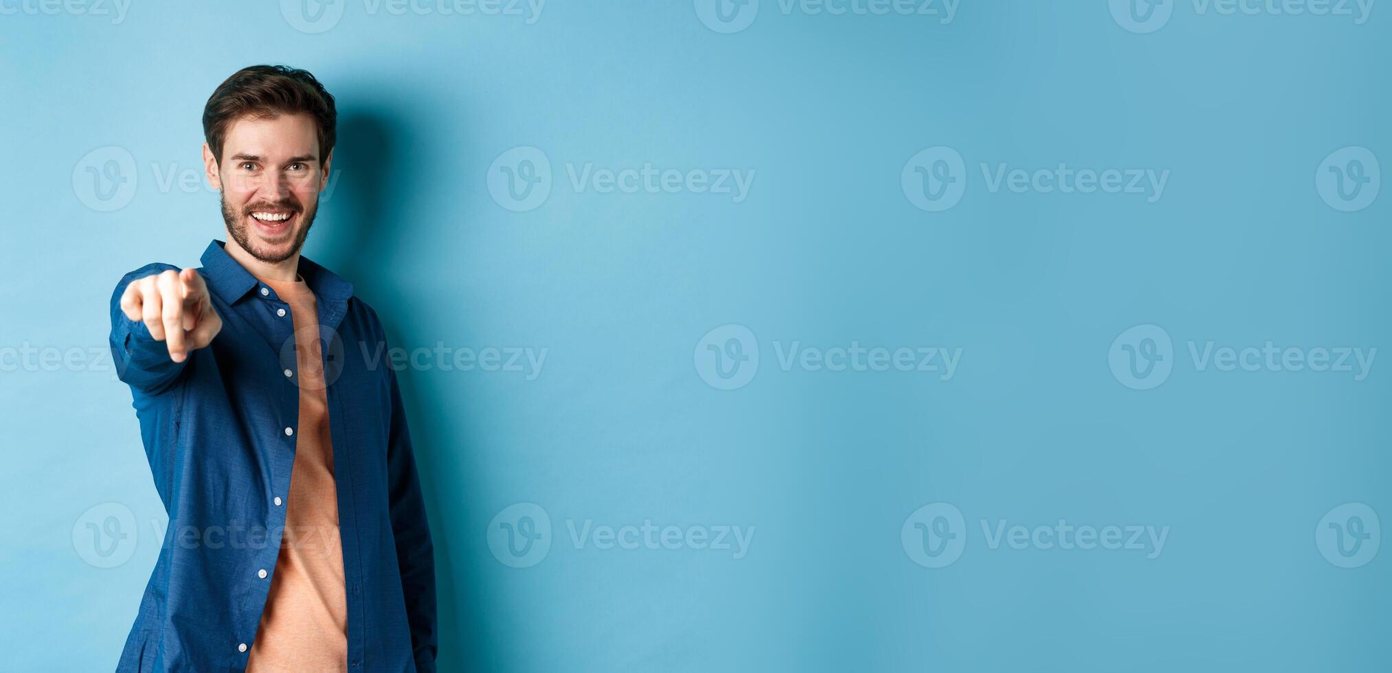 Confident smiling guy choosing or inviting you, pointing finger at camera decisive, standing on blue background photo
