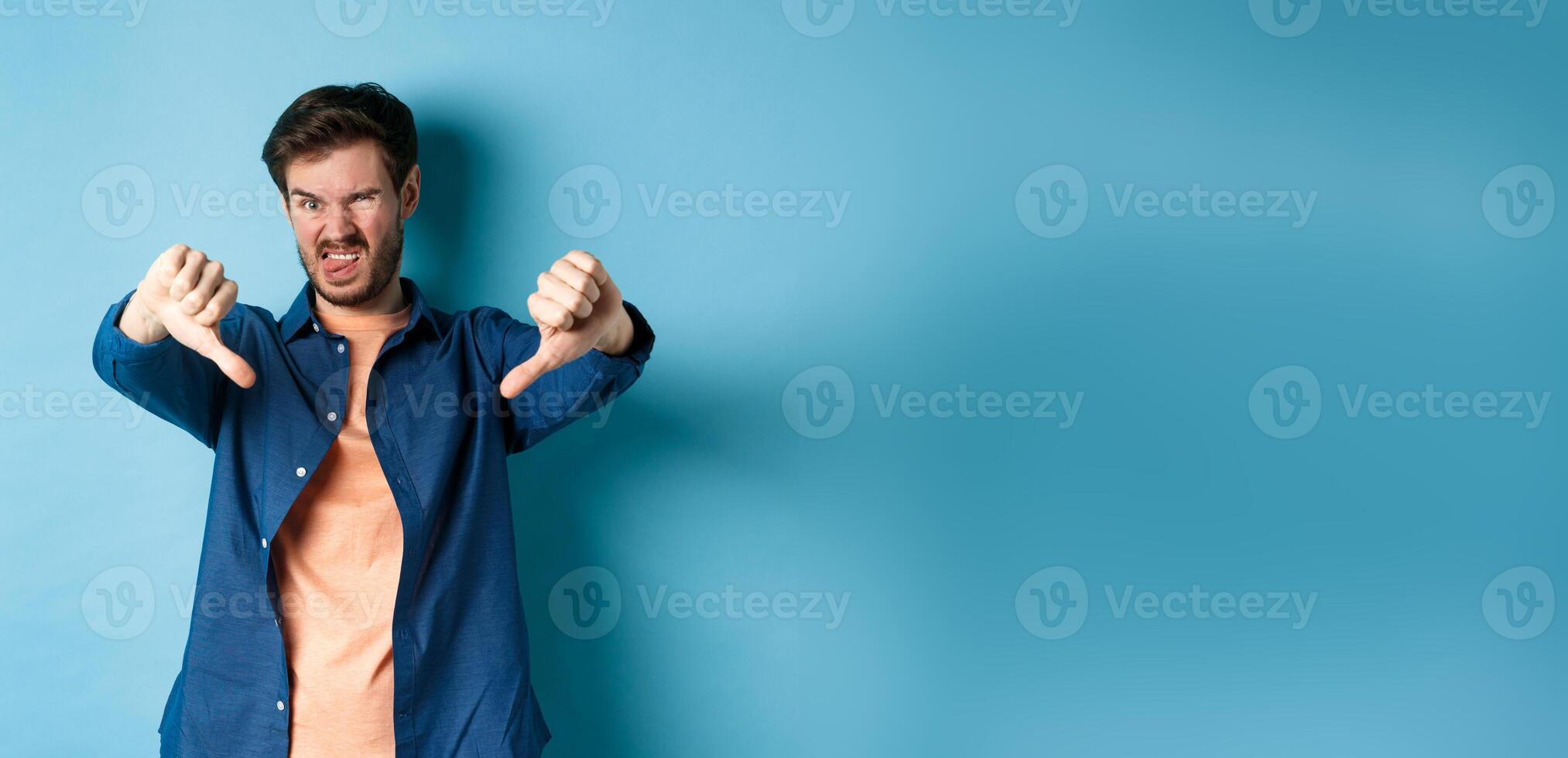 Disgusted guy express negative opinion, showing thumbs down and tongue, frowning upset, disapprove something bad, standing on blue background photo