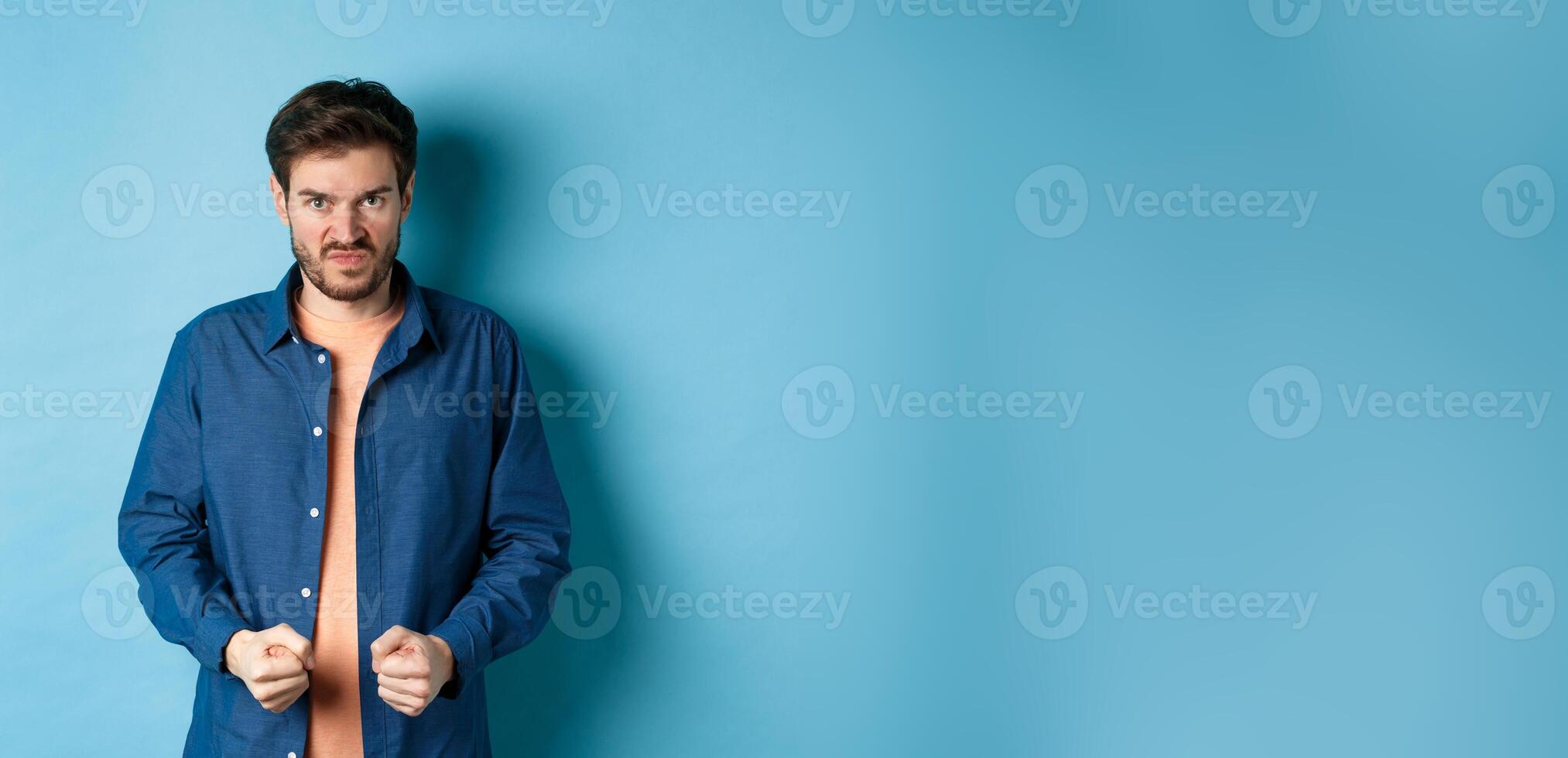 Angry and pissed-off man clench fists and stare with disdain at camera, trying to keep himself together, look with rage, standing on blue background photo