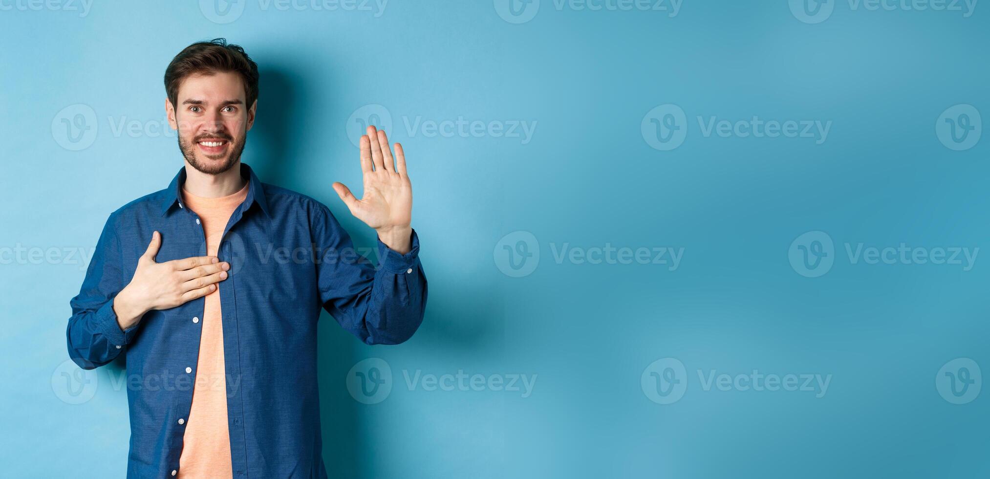 Honest young man smiling and making promise, holding hand on heart and arm raised, pledge or give oath, swearing to tell truth, standing on blue background photo