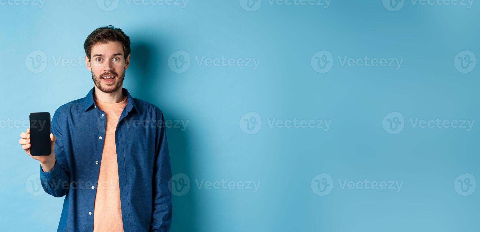 Happy caucasian guy showing empty mobile screen and smiling, standing on blue background photo
