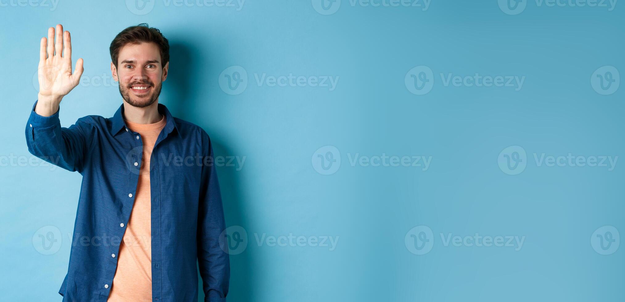 Friendly young man smiling and raising hand up for high five, waiving to greet or say hello, standing on blue background photo