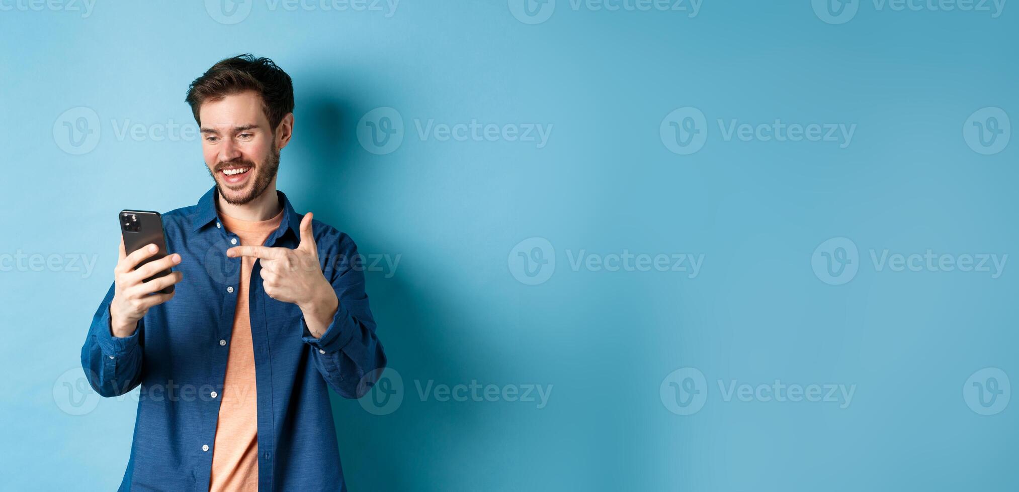 Image of handsome smiling man reading mobile screen and pointing at phone, checking out online promo or smartphone app, standing on blue background photo