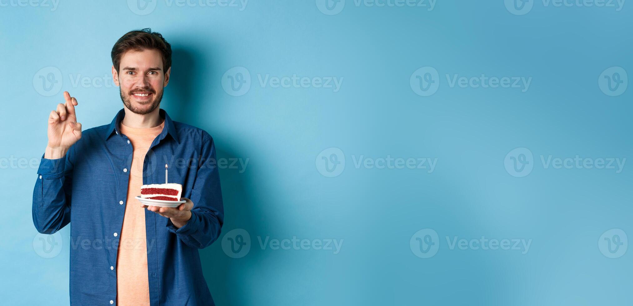 Celebration and holiday concept. Hopeful young man making wish on birthday cake, cross fingers for good luck and smiling positive, standing on blue background photo