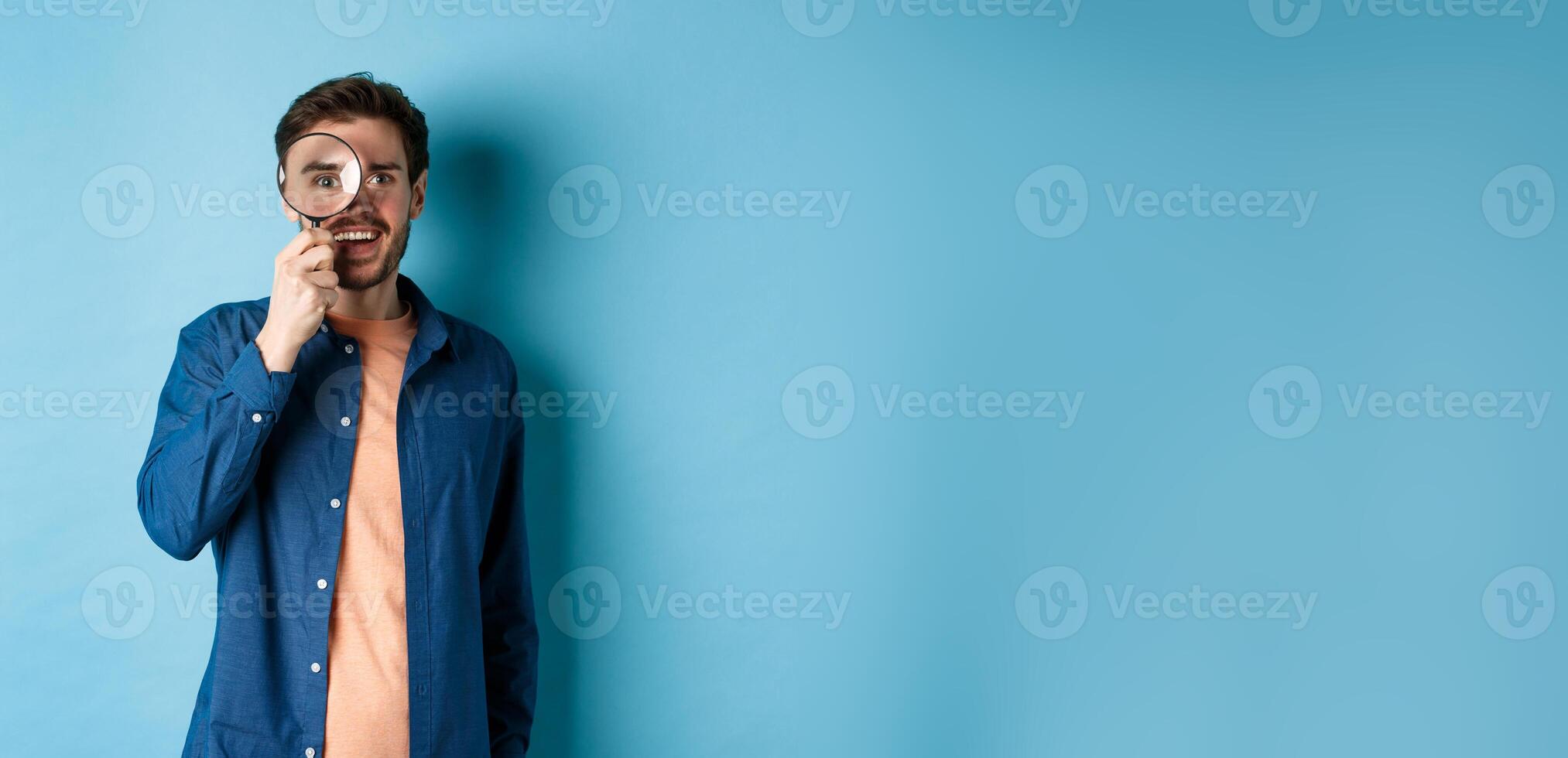 Cheerful young man found something good, smiling and looking through magnifying glass, standing on blue background photo