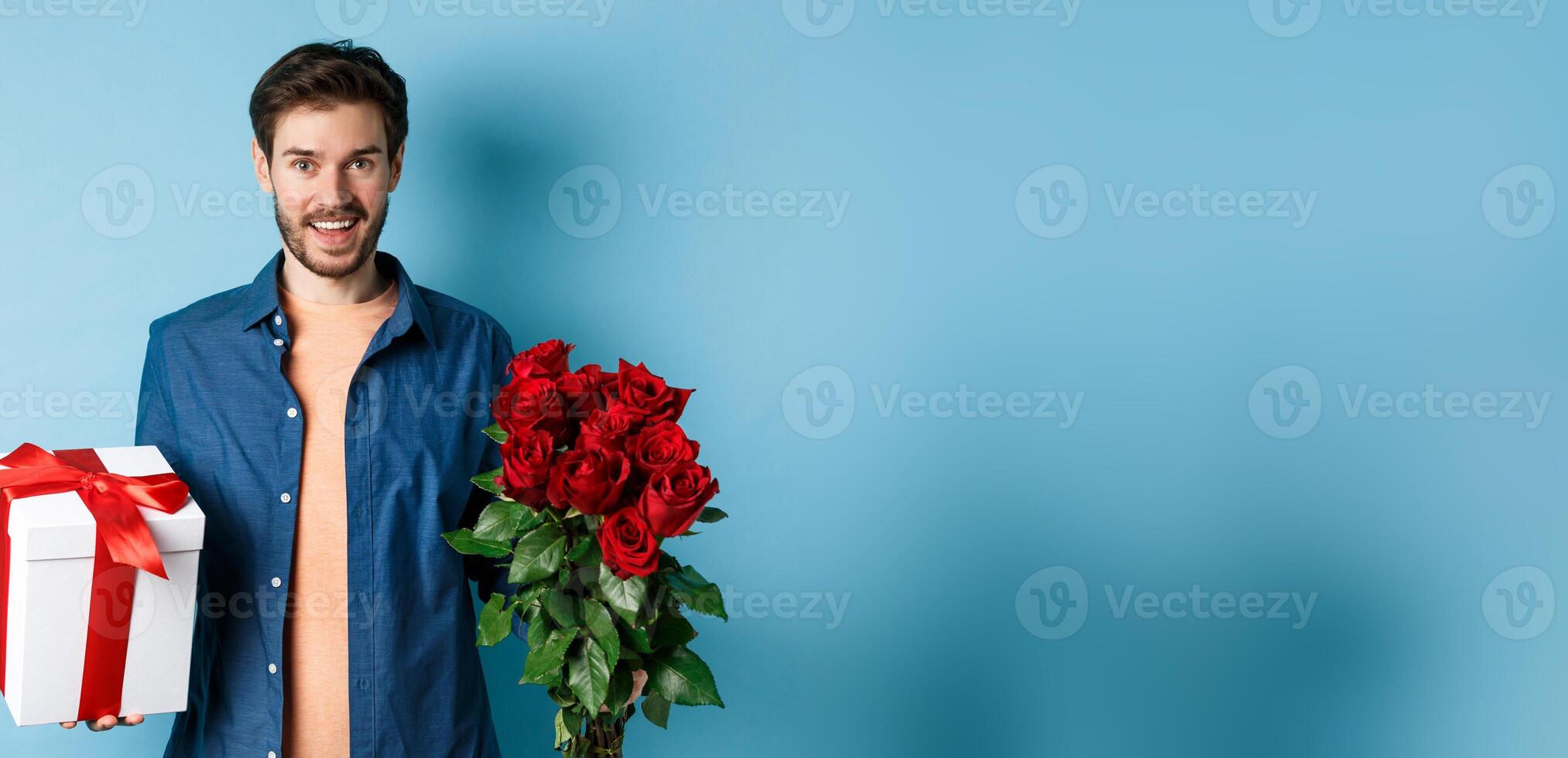 Love and relationship concept. Happy young man bring flowers and gift on romantic date. Boyfriend wish bouquet of roses and present standing on blue background photo