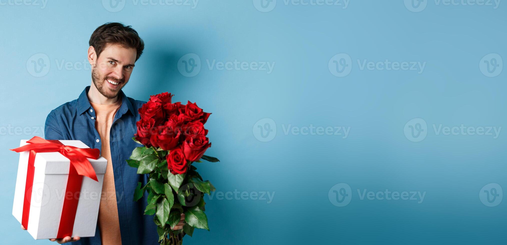 Love and Valentines day concept. Romantic smiling man giving you gift box and bouquet of flowers on date, standing over blue background photo