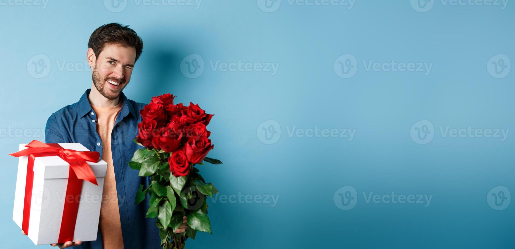 Love and Valentines day concept. Happy guy winking and smiling at camera, presenting a gift and bouquet of flowers to girlfriend, standing over blue background photo