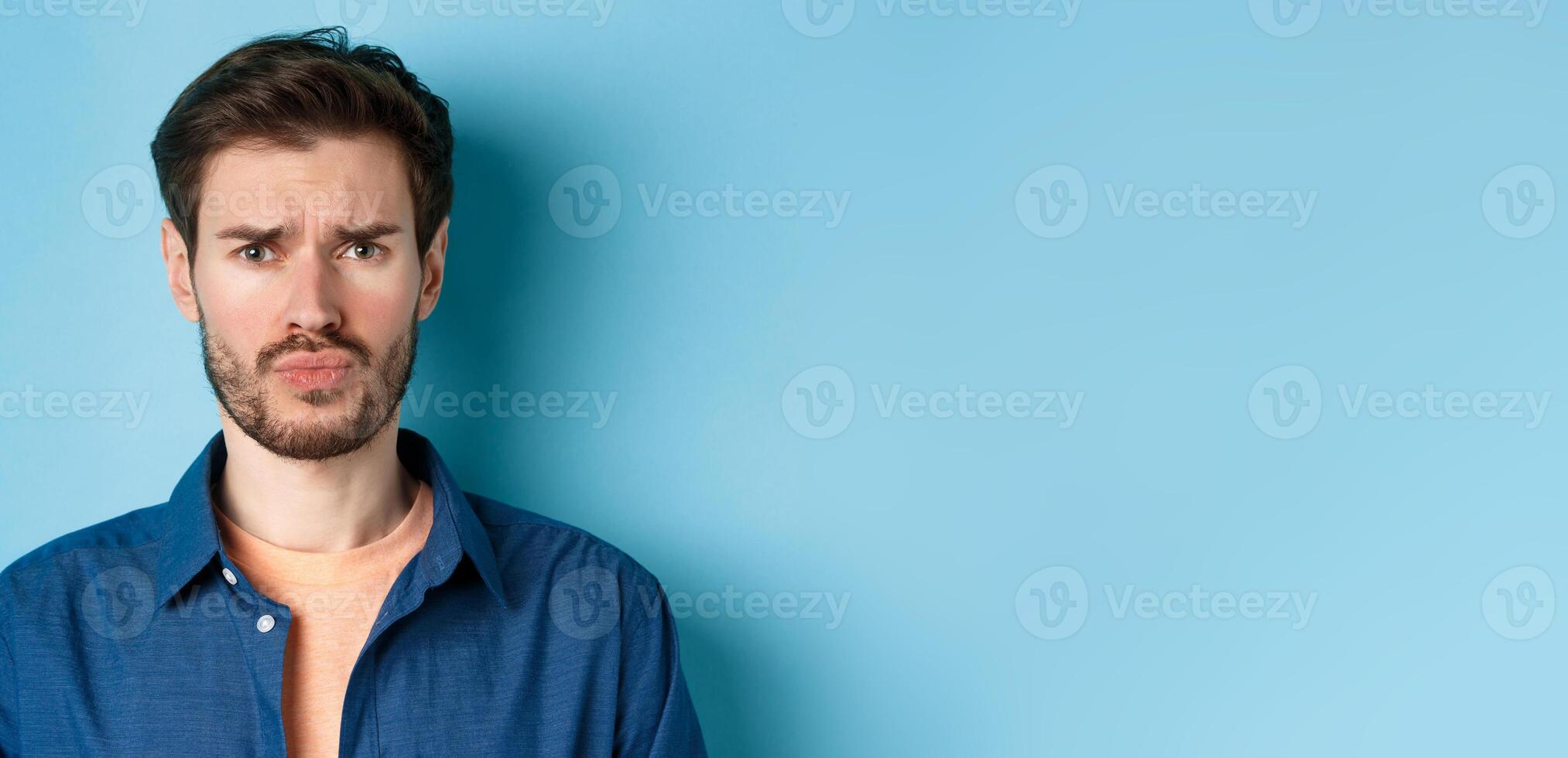 Close up of sad and offended young man sulking at someone, frowning upset, standing on blue background photo