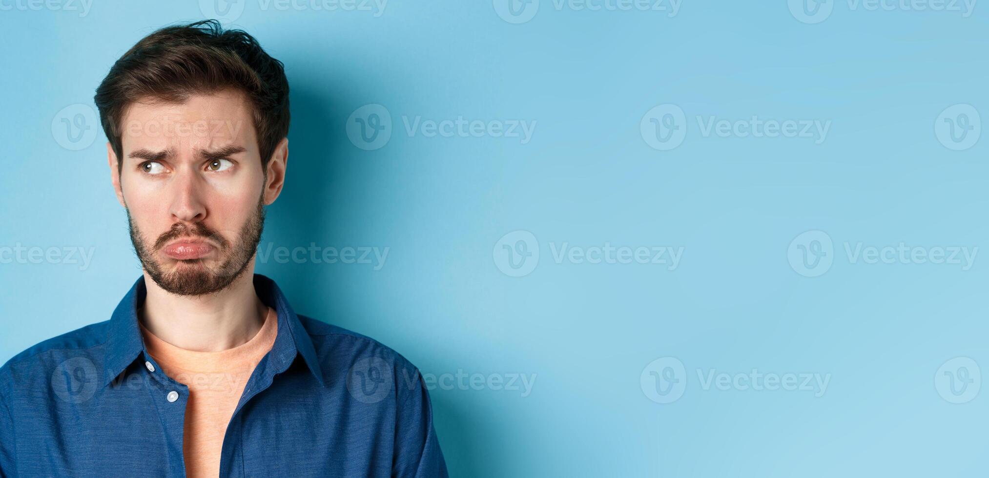Miserable and disappointed guy acting childish, sulking and looking left at empty space with offended face, standing on blue background photo