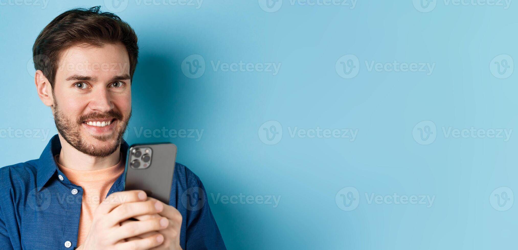 Close up of young guy with beard, smiling happy at camera and holding mobile phone, standing on blue background photo
