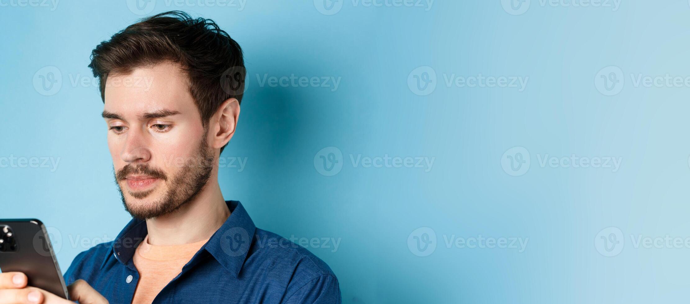 Close up of handsome bearded man reading mobile phone message, looking at screen serious, standing on blue background photo