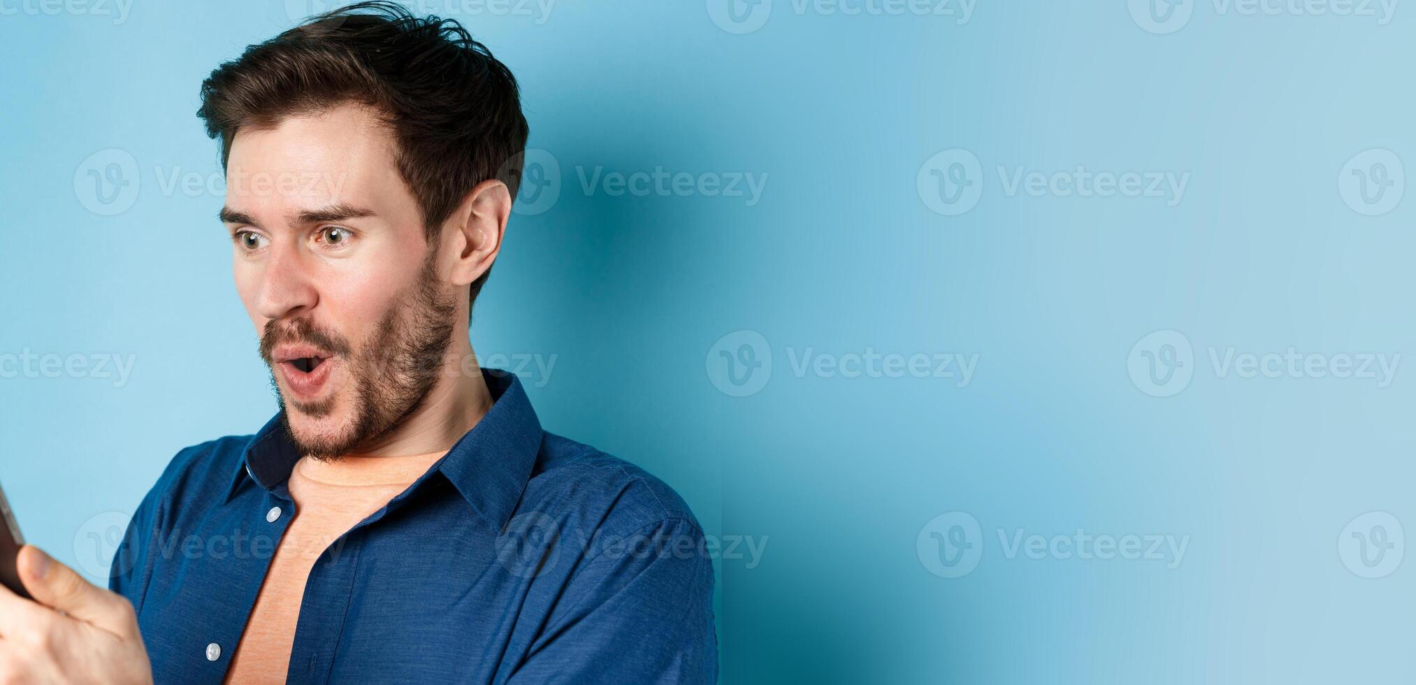 Image of happy young man stare at mobile screen, winning online and cheering, standing on blue background photo