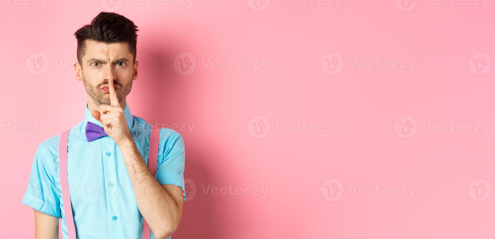 Angry and grumpy man in funny bow-tie shushing at camera, frowning and demand silence, tell to be quiet, scolding someone loud, standing on pink background photo