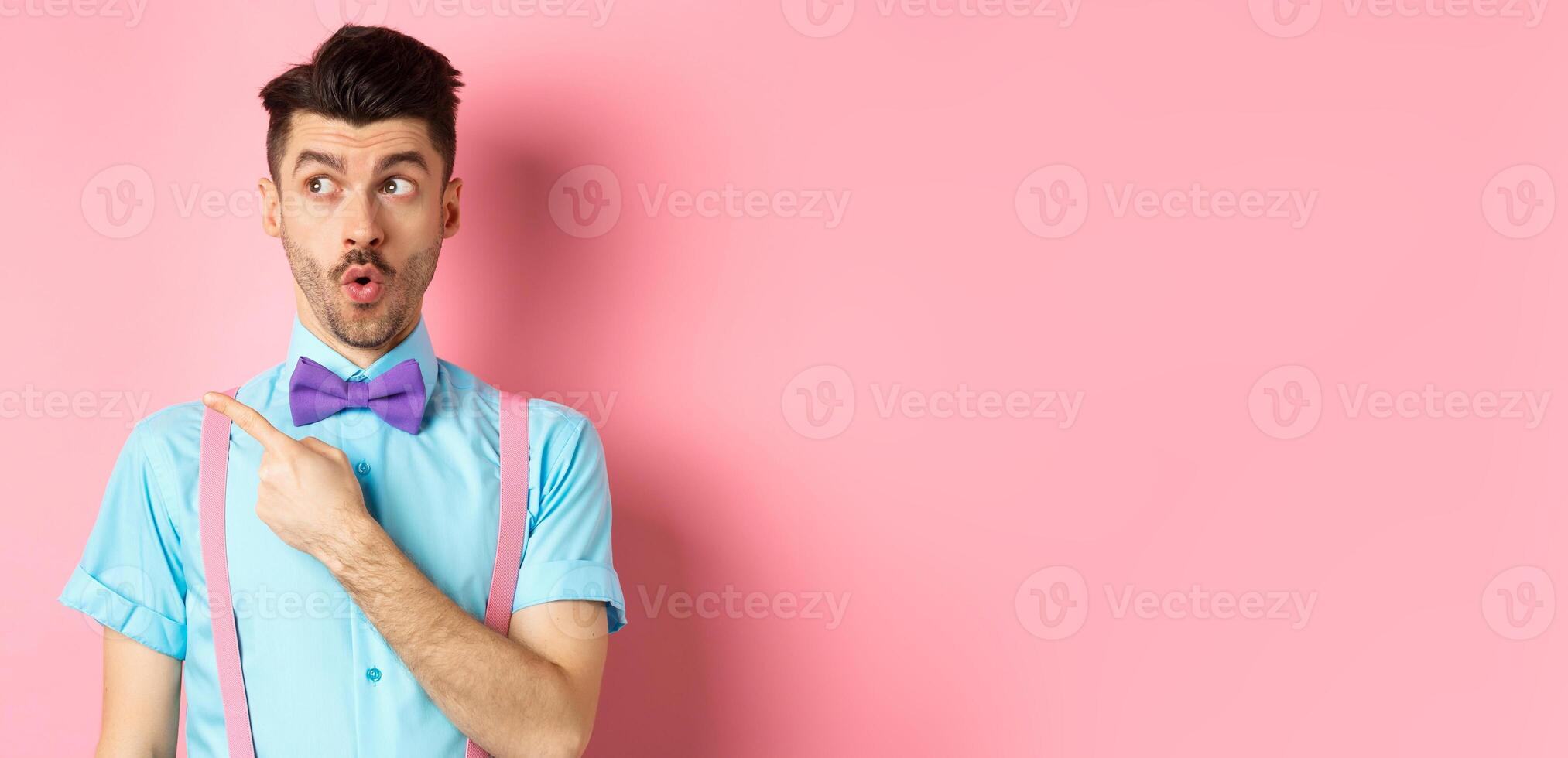 Intrigued young man pointing finger left at logo, checking out promo offer, standing in bow-tie and classy outfit, pink background photo