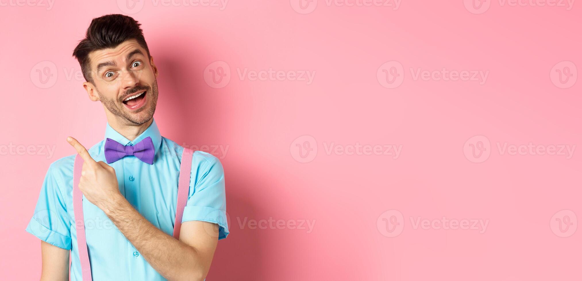 Funny male model in bow-tie, tilt head and pointing finger left, inviting to look, check it out gesture, showing advertisement on pink background photo