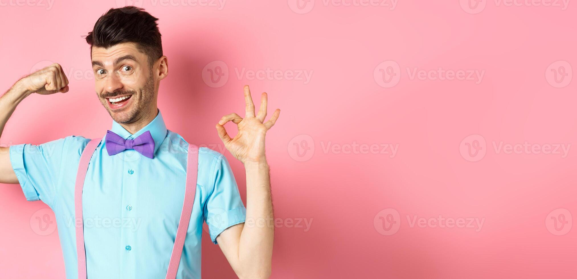 Handsome young man with moustache and bow-tie, flex biceps, showing strong arm and okay sign, workout at gym, standing on pink background photo