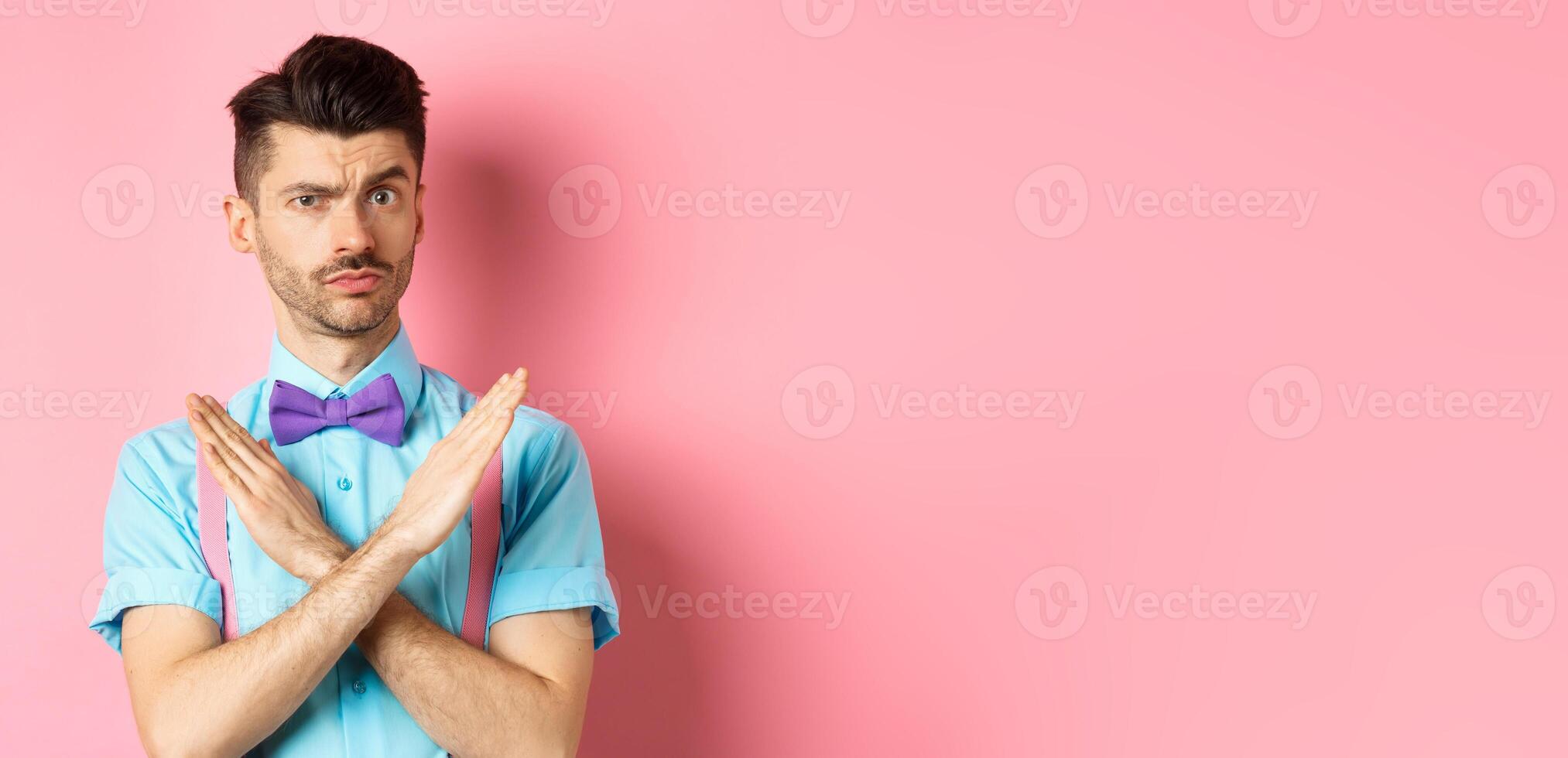 Serious-looking skeptical guy showing cross sign to stop, prohibit something bad, forbid action, standing on pink background photo