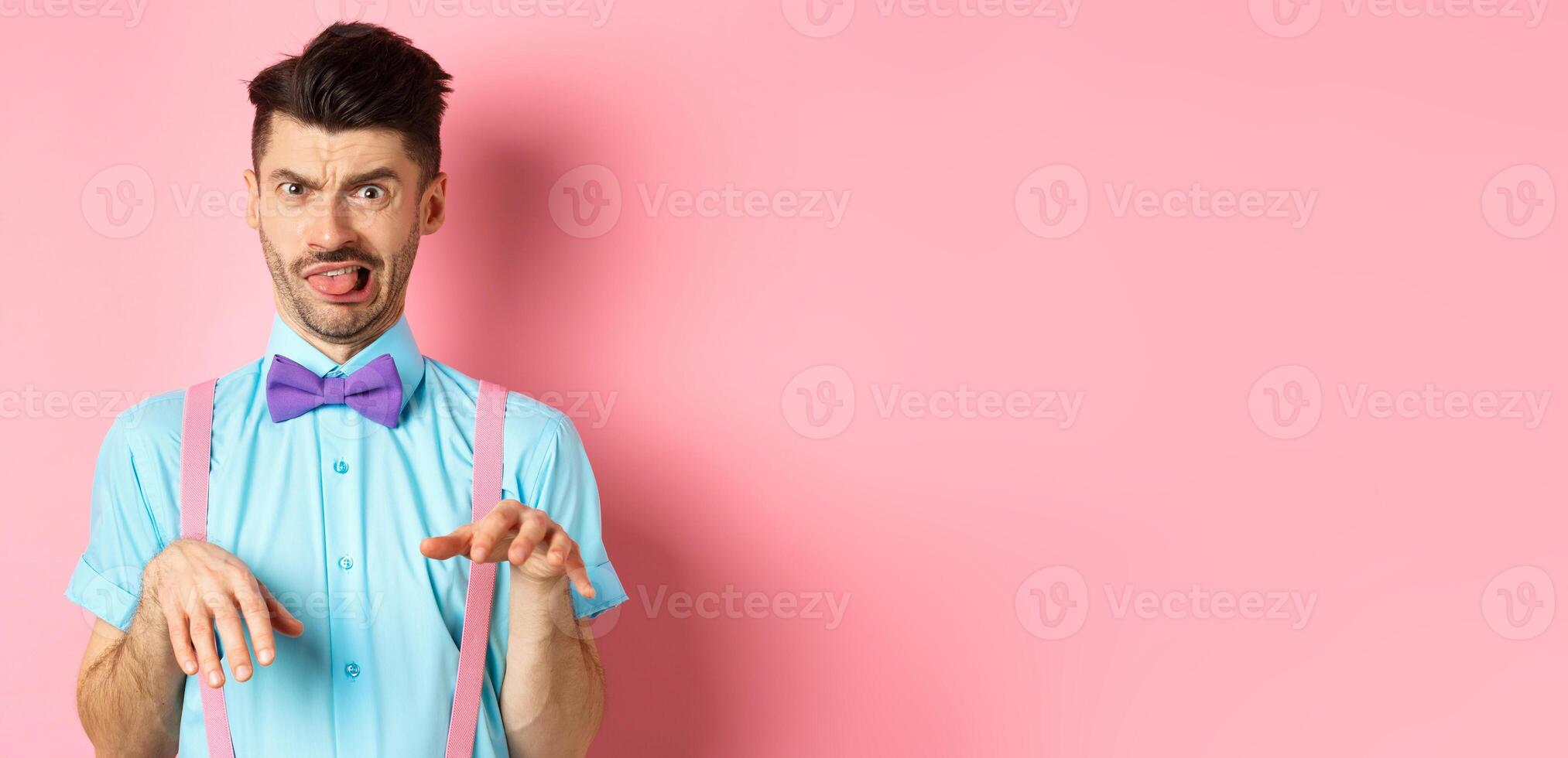 Funny guy looking at something disgusting with aversion and cringe, show tongue and shaking hands in disapproval, standing on pink background photo