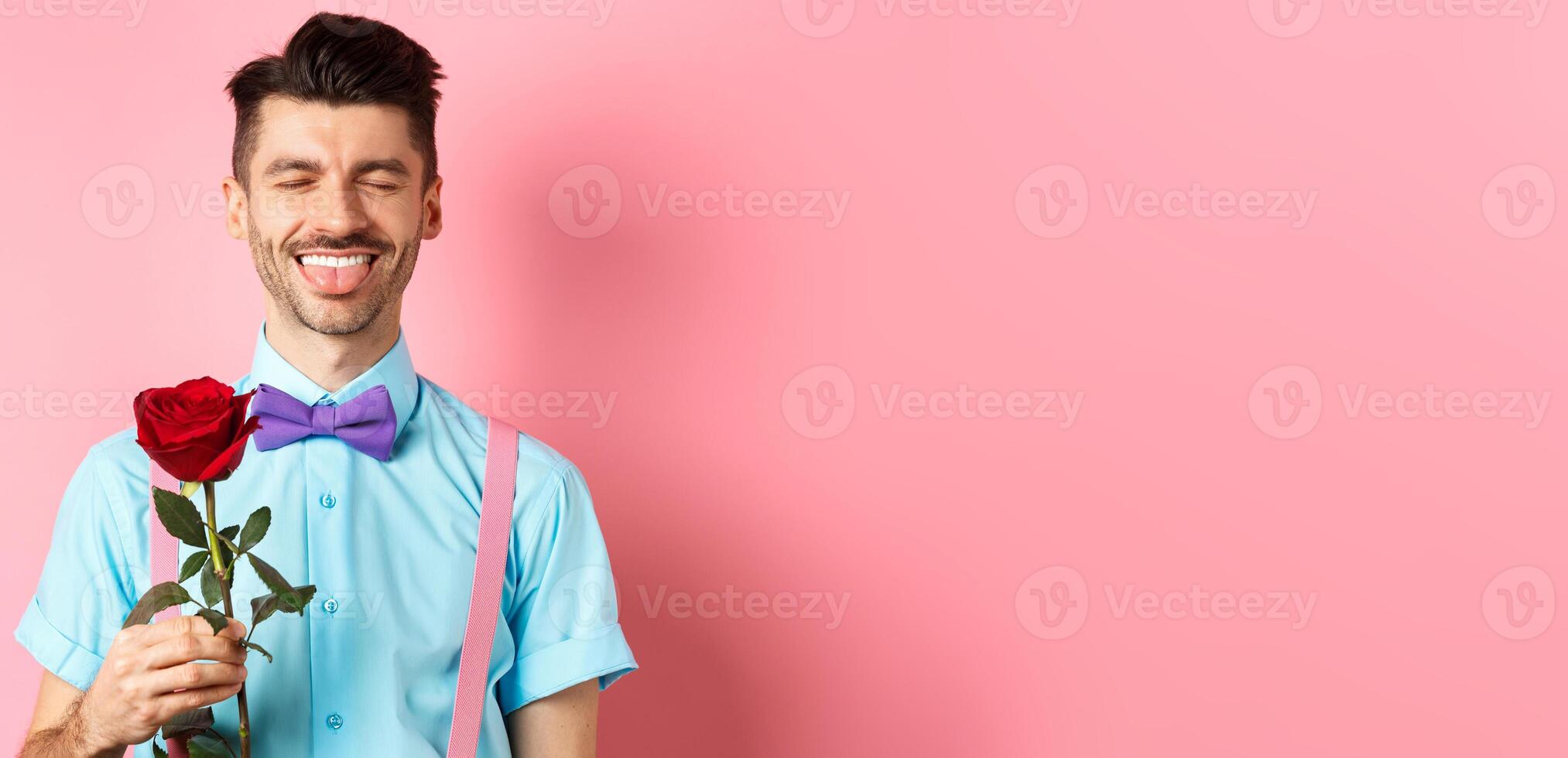 Happy man showing tongue and smiling, holding red rose for girlfriend on Valentines day, enjoying romantic date, pink background photo