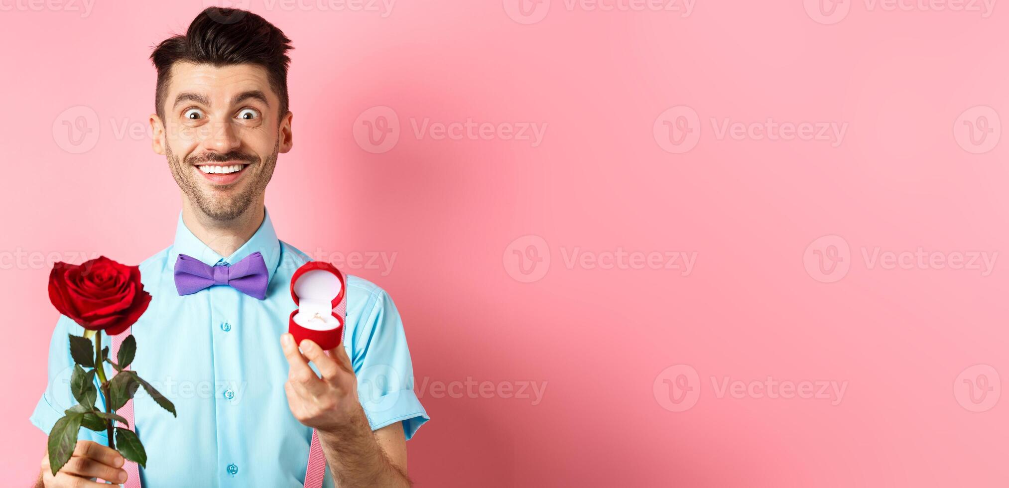 Valentines day. Smiling handsome man asking to marry him, showing engagement ring and red rose, standing romantic on pink background photo