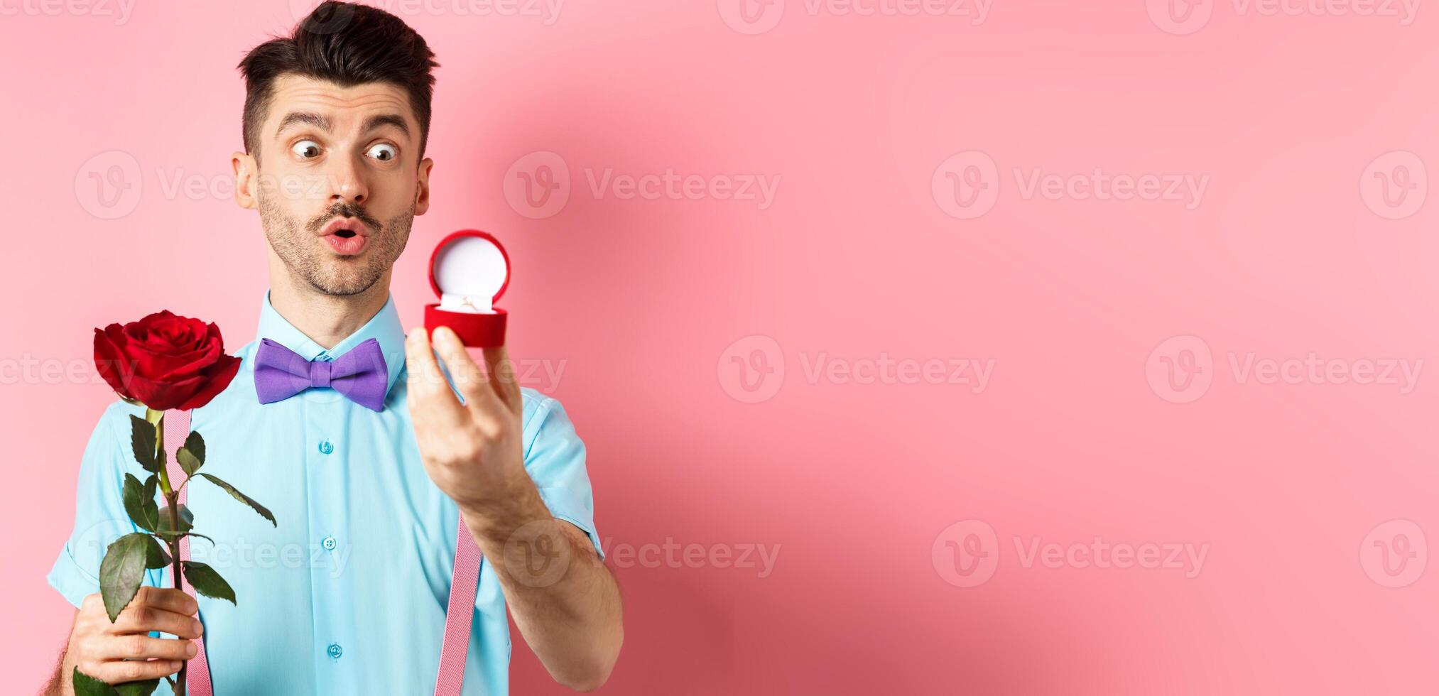 Valentines day. Romantic funny guy going to make wedding proposal, asking to marry him, holding red rose and looking at engagement ring with excitement, pink background photo