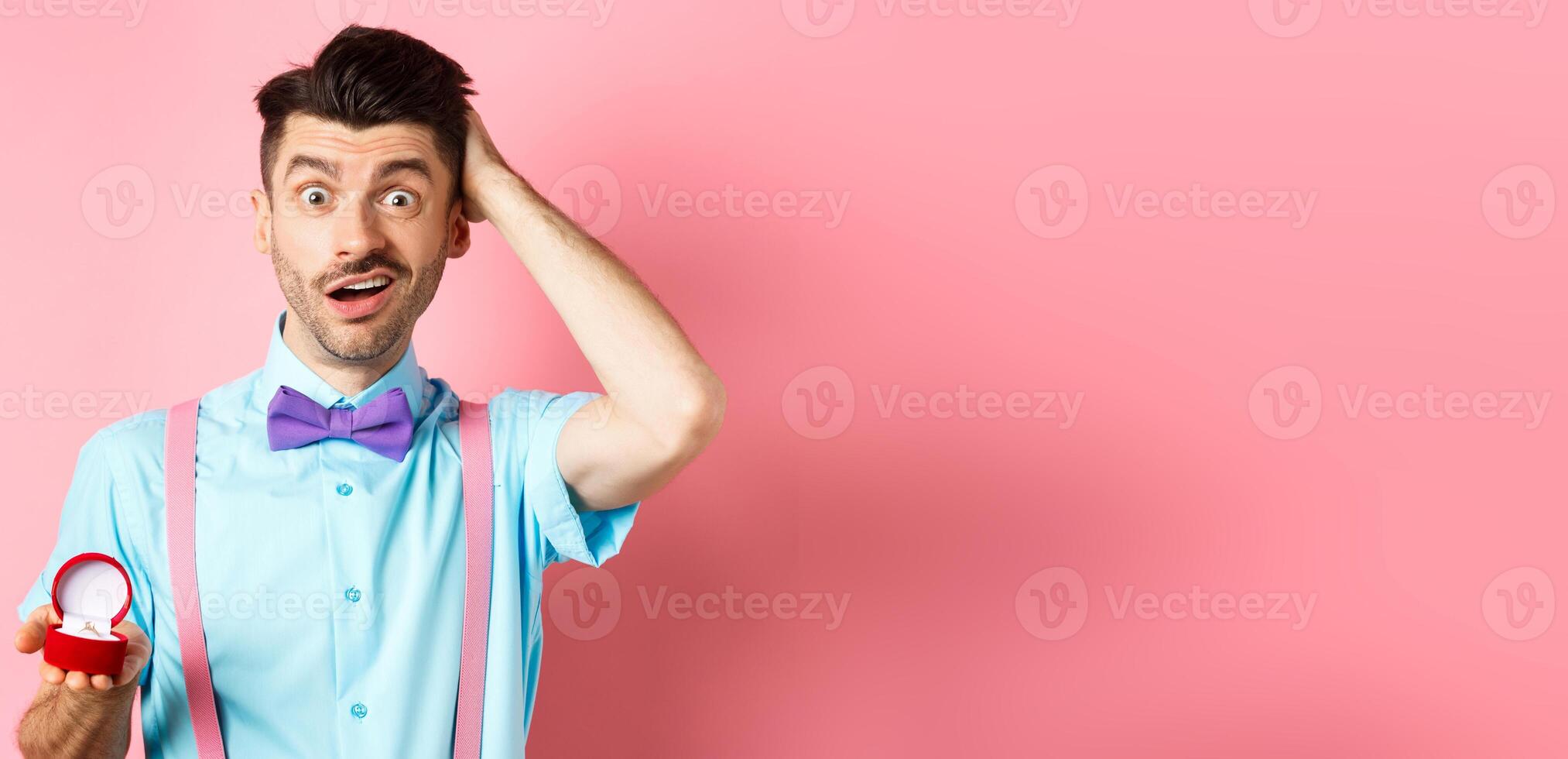 Valentines day. Confused funny guy standing with engagement ring, scratching head with unsure face, dont know what say, making proposal, standing over pink background photo