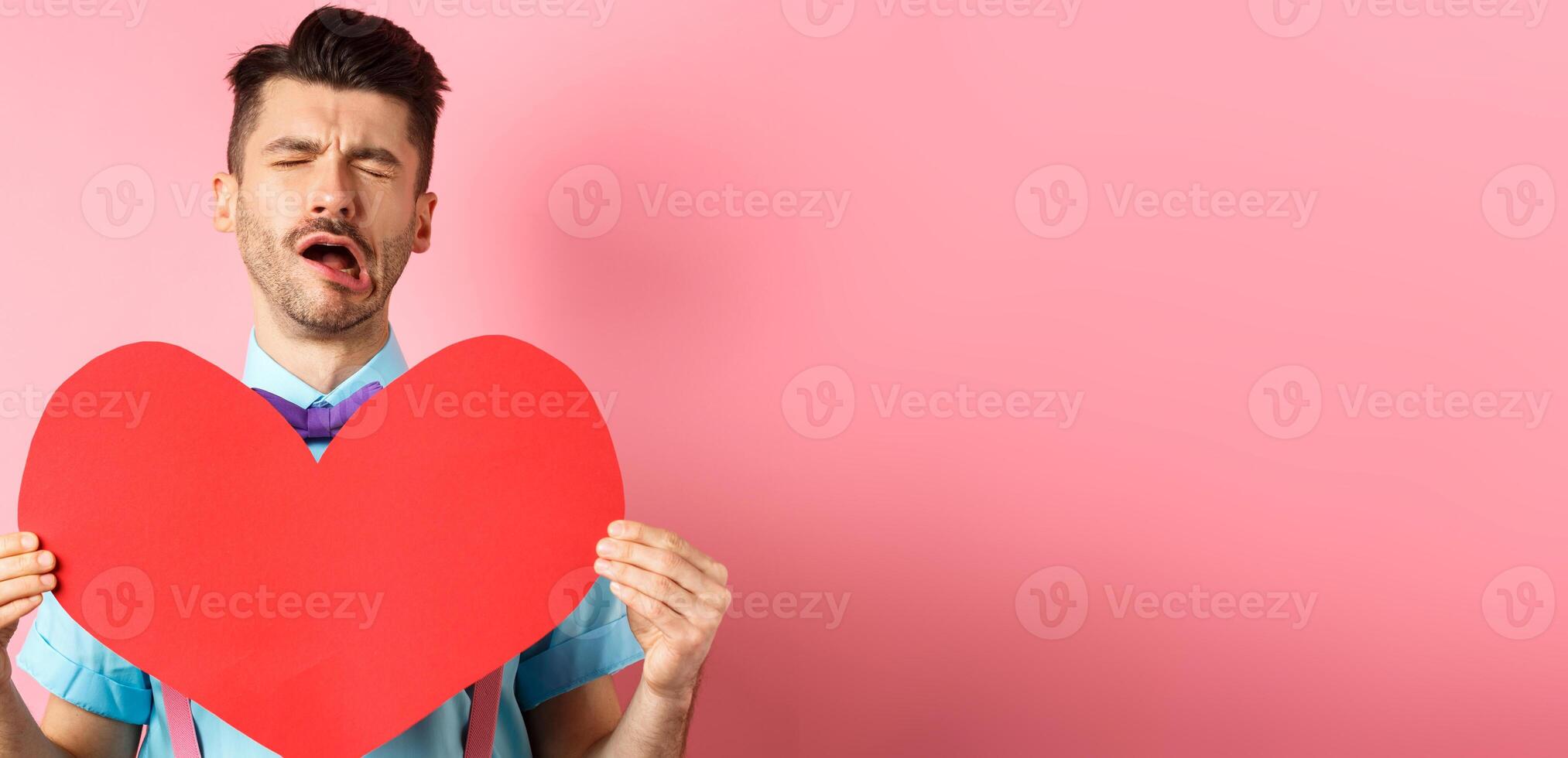 Valentines day concept. Sad and lonely man feeling heartbroken, being rejected, showing big red heart cutout and crying from break-up, standing on pink background photo
