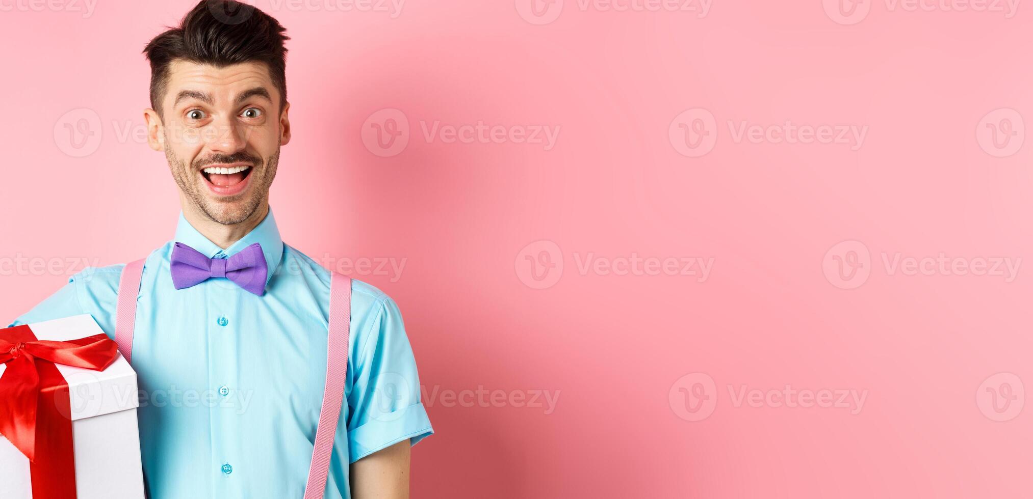 Holidays and celebration concept. Happy young man in festive outfit holding big present box for birthday, standing cheerful and smiling, pink background photo