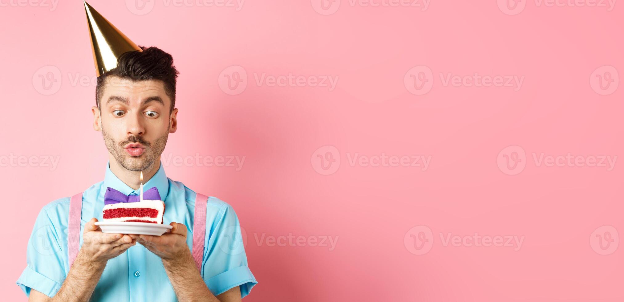 Holidays and celebration concept. Happy young man in birthday party hat blowing candle on cake, making wish on b-day, standing over pink background photo