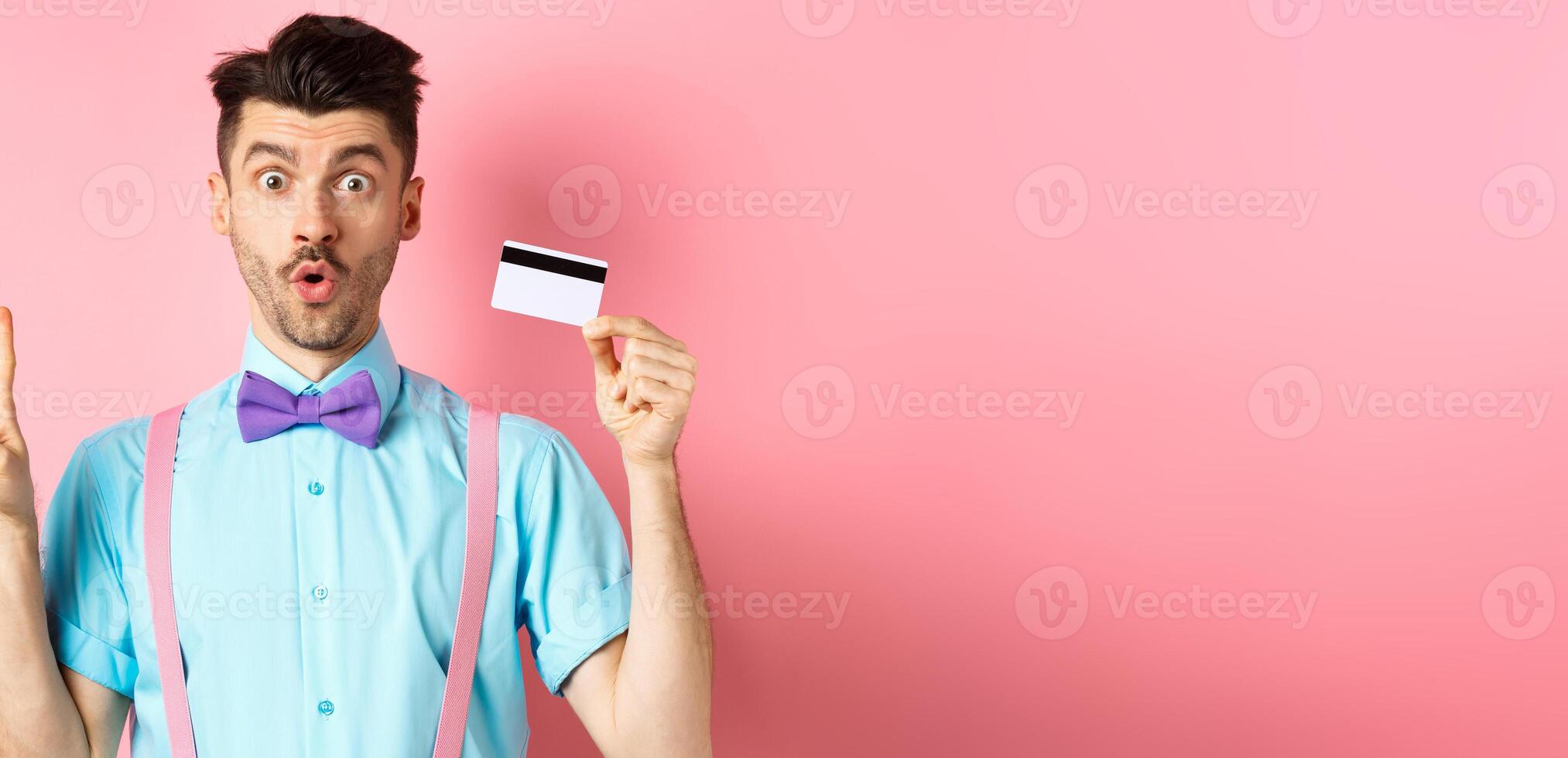 Shopping concept. Image of funny guy looking surprised while showing plastic credit card and victory sign, checking out promo deal, standing on pink background photo