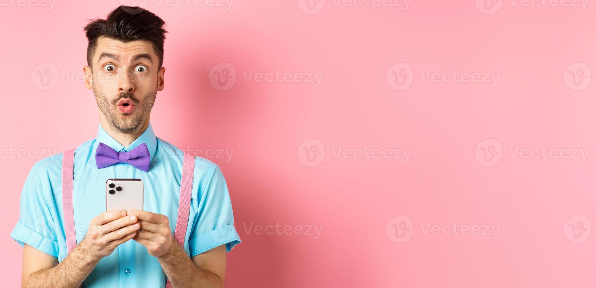 Online shopping. Intrigued guy in bow-tie, checking out internet promo offer on mobile phone, say wow at camera, holding smartphone, standing on pink background photo