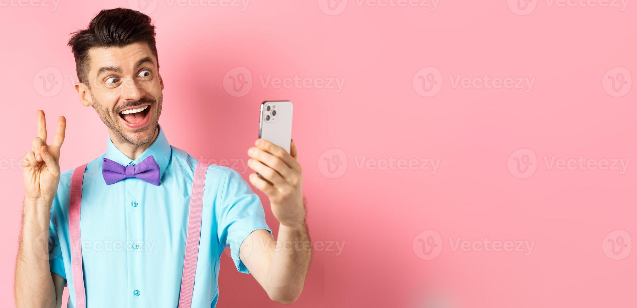 Technology concept. Funny man with moustache and bow-tie taking selfie on smartphone, showing peace sign and smiling at mobile camera, pink background photo