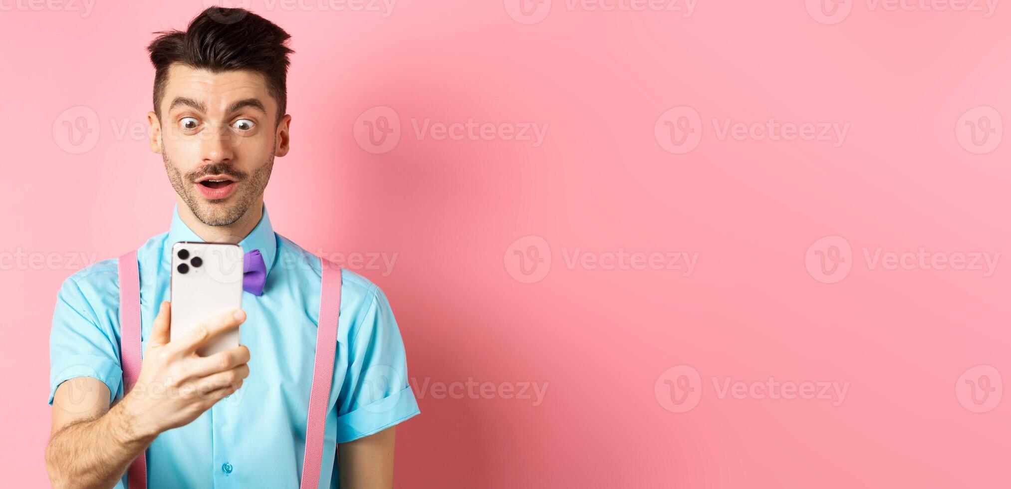 Impressed man reading mobile screen with amazed face, seeing online promo offer, standing over pink background photo