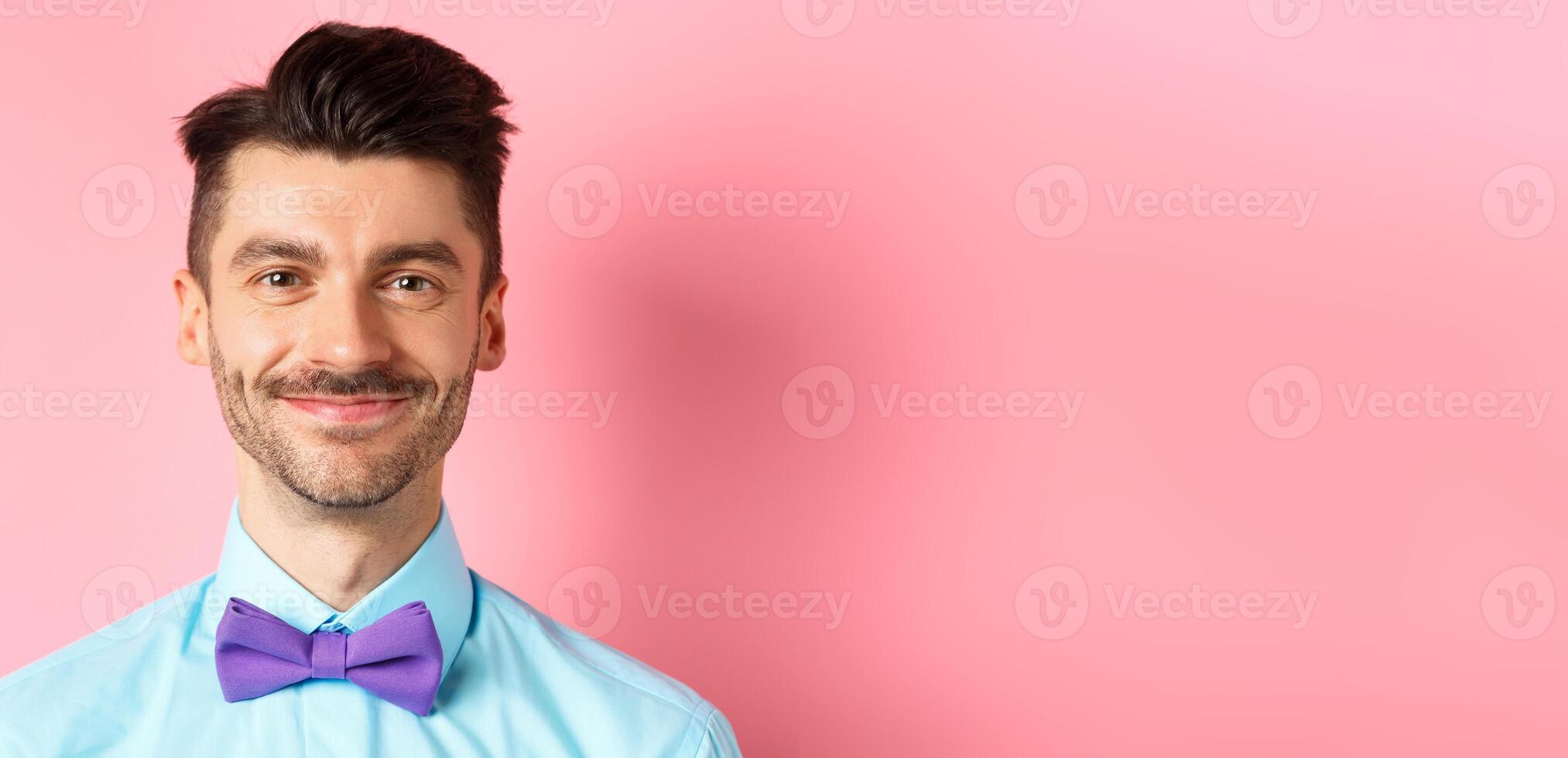 Close-up of smiling hansome man with moustache, wearing bow-tie and shirt, standing cheerful on pink background photo